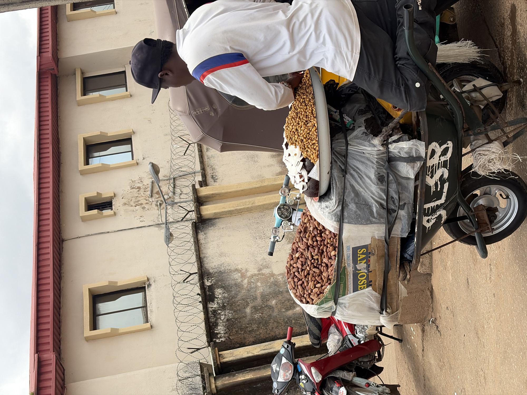 In Jos, Nigeria, a street vendor is depicted from the side, wearing a black baseball cap and a white shirt with red and blue accents on the sleeves. He is seated on a loaded wheelbarrow, which is positioned next to a motorcycle. The wheelbarrow is stacked with various items, including a large bag of what appears to be dates, several other bags of food items covered in plastic, and a large metal tray filled with roasted nuts and some white pieces of food. The vendor is reaching into the tray, seemingly serving a customer who is out of frame. The scene is set against a building with a red corrugated roof and beige walls, featuring several windows with unique angled frames and barbed wire fencing along the front. The ground is paved with what appears to be asphalt or concrete, with some debris scattered around. The sky is overcast.