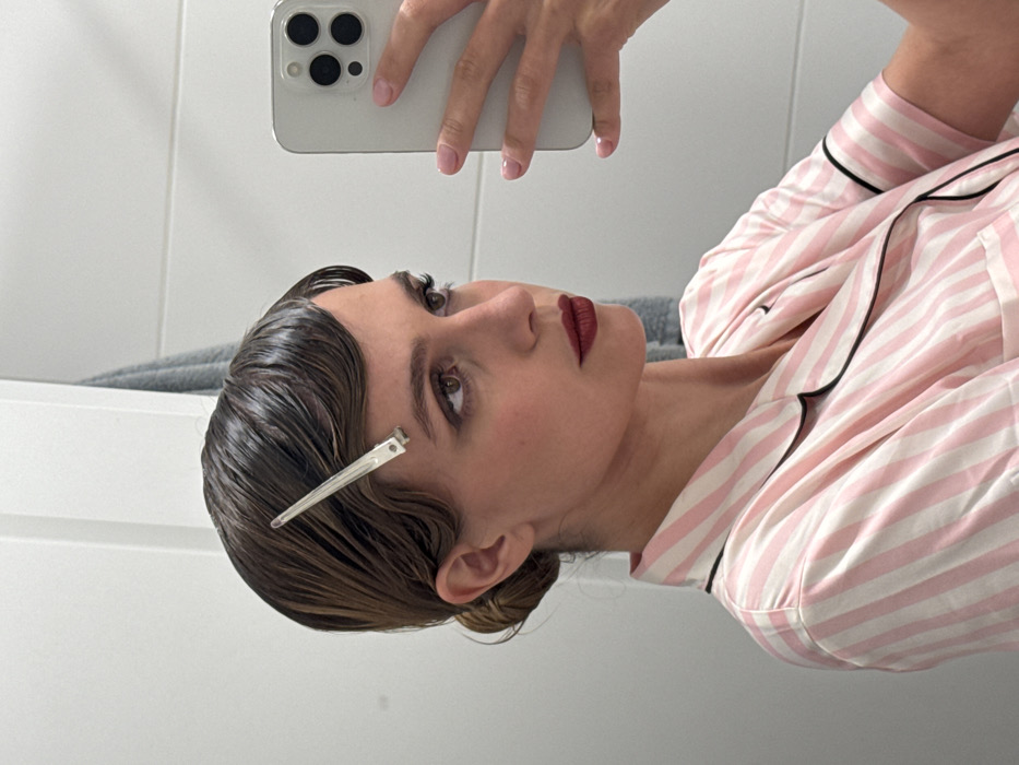 Location: Indoors, likely a bathroom or dressing area within a private residence in Unknown, Unknown.
Scene Description: A female-presenting individual is depicted in a close-up, chest-up selfie captured via a mirror reflection. The individual faces right, her gaze directed slightly upward. Her dark brown hair is styled in distinct finger waves across her forehead and temples, appearing wet or gelled, and secured with a silver alligator clip above her right temple. The remainder of her hair is pulled back into a low bun. She exhibits light skin, dark eyes accentuated with dark eyeliner and mascara, defined eyebrows, and dark red lipstick. She wears a light pink and white striped pajama top with black piping along the collar and short sleeve edges. Her left hand, visible in the reflection, holds a light-colored smartphone with a prominent triple-lens camera system, indicating the selfie-taking action. Her fingernails are manicured with light pink polish. The background consists of a plain white wall, likely tiled, and a grey towel draped in the upper right quadrant.