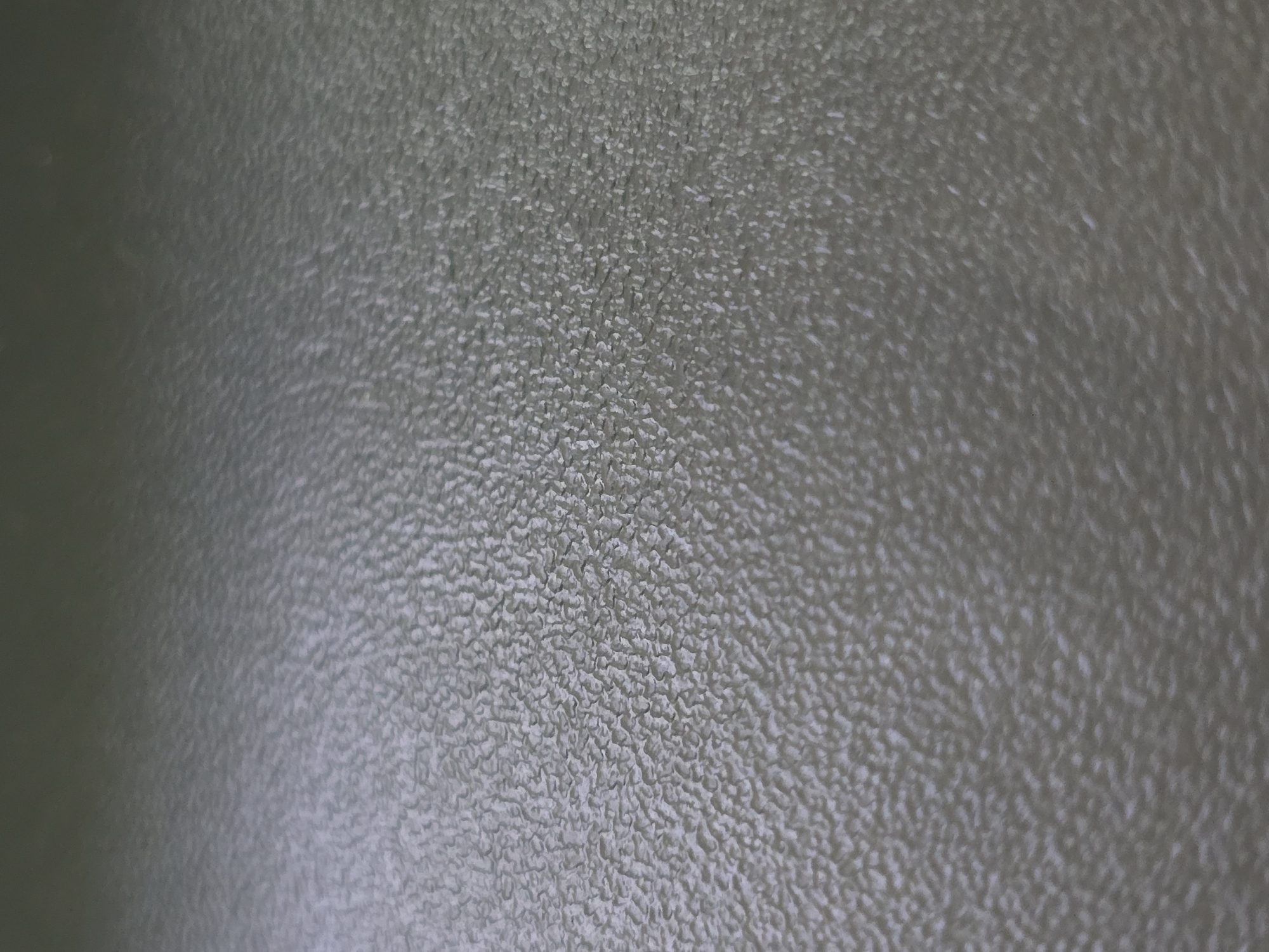The image shows a close-up, abstract view of a textured surface. The texture is granular and bumpy, with an uneven pattern of raised and recessed areas. The color is a muted, desaturated greenish-gray, with subtle variations in shade that suggest a slight sheen or subtle lighting. The focus is shallow, with the foreground appearing sharper than the background, which gradually blurs. No specific objects, people, or actions are discernible. The image does not provide any information about its location in the city of Kotiv, Ukraine.
