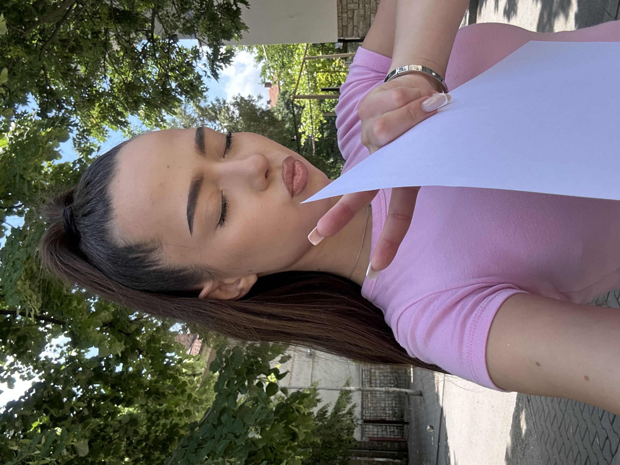 A young woman is depicted taking a selfie outdoors, holding a plain white piece of paper. She has long, dark brown hair pulled back into a high ponytail. Her eyes are closed, and her lips are slightly pursed, adorned with a glossy, nude-colored lipstick, conveying a playful expression. She is wearing a light pink short-sleeved top and a silver-toned bracelet on her left wrist. Her fingernails are neatly manicured in a French tip style.
The background is dominated by lush green trees with dense foliage, suggesting an outdoor setting like a park or a tree-lined area. Glimpses of a bright blue sky with scattered clouds are visible through the leaves. In the mid-ground, there are hints of a building with a textured stone or brick facade. A paved surface, possibly a sidewalk or road, is visible in the lower right corner. The bright lighting indicates it is daytime with clear, sunny weather. There is no legible text visible on the paper or elsewhere in the image.