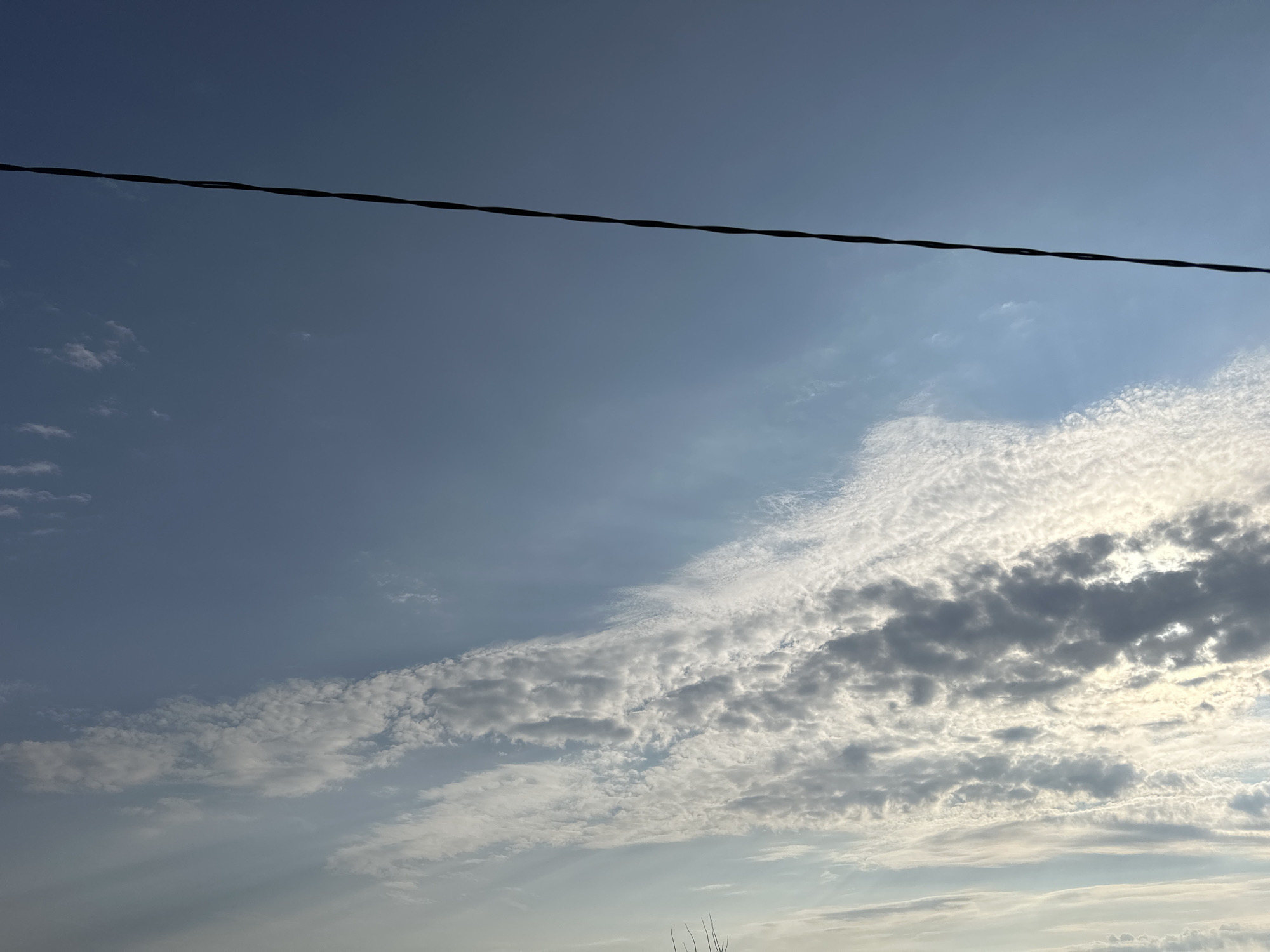A thin, dark power line bisects the upper portion of the frame horizontally, stretching across a pale blue sky. Wispy white clouds, tinged with grey in places, begin to gather in the lower right quadrant, becoming more dense and textured as they extend towards the center. The lower portion of the image is a gradient of lighter sky, with some faint cloud formations visible. The scene is devoid of people or specific landmarks, suggesting an open-air environment. Given the location provided, the image likely depicts a sky view over the city of Ulyanovsk, Russia.