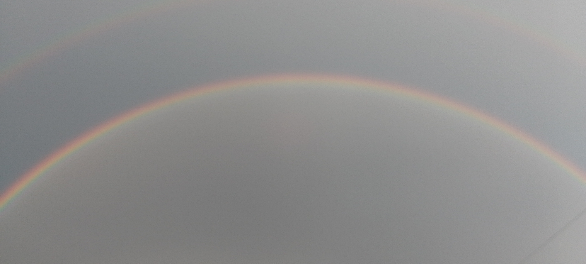The image shows a faint double rainbow against a gray sky. The primary rainbow is more visible, displaying a curved arc of muted colors, predominantly yellow, orange, and red. A secondary, fainter rainbow is visible above and parallel to the main arc, with its colors appearing in reverse order. The overall impression is of a soft, atmospheric phenomenon. There are no people, buildings, or specific landmarks visible to indicate a location in Cherkasy, Ukraine, or any other city. The environment is purely sky.