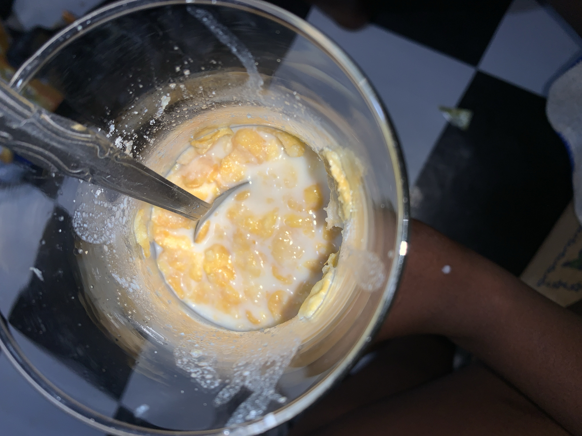 An overhead shot shows a clear glass filled with cornflakes and milk. A metal spoon is in the glass, partially submerged in the milk and cereal. The glass has milk residue clinging to the interior sides. The background reveals a black and white checkered surface and a person's arm and leg. The photo's origin is uncertain, and the location of Rafin Par, Nigeria, cannot be confirmed from the image alone.