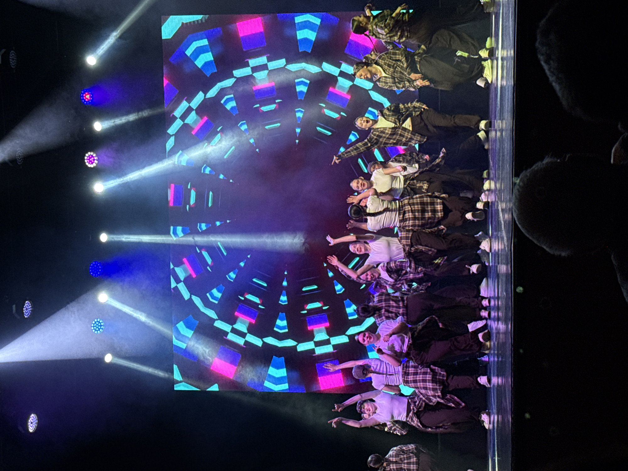 The image depicts a group of dancers performing on a stage in what appears to be an indoor venue. The dancers are arranged in a line across the stage. They are a mix of genders and are wearing a combination of white t-shirts and plaid shirts with dark-colored pants. Behind them is a large screen displaying an abstract geometric pattern in shades of blue, cyan, and pink. Stage lighting is visible, with several spotlights shining on the dancers from the left. The atmosphere suggests a performance setting with a focus on choreography and visual effects. This photo may be taken during an event in the city of Chaikovsky, Russia.