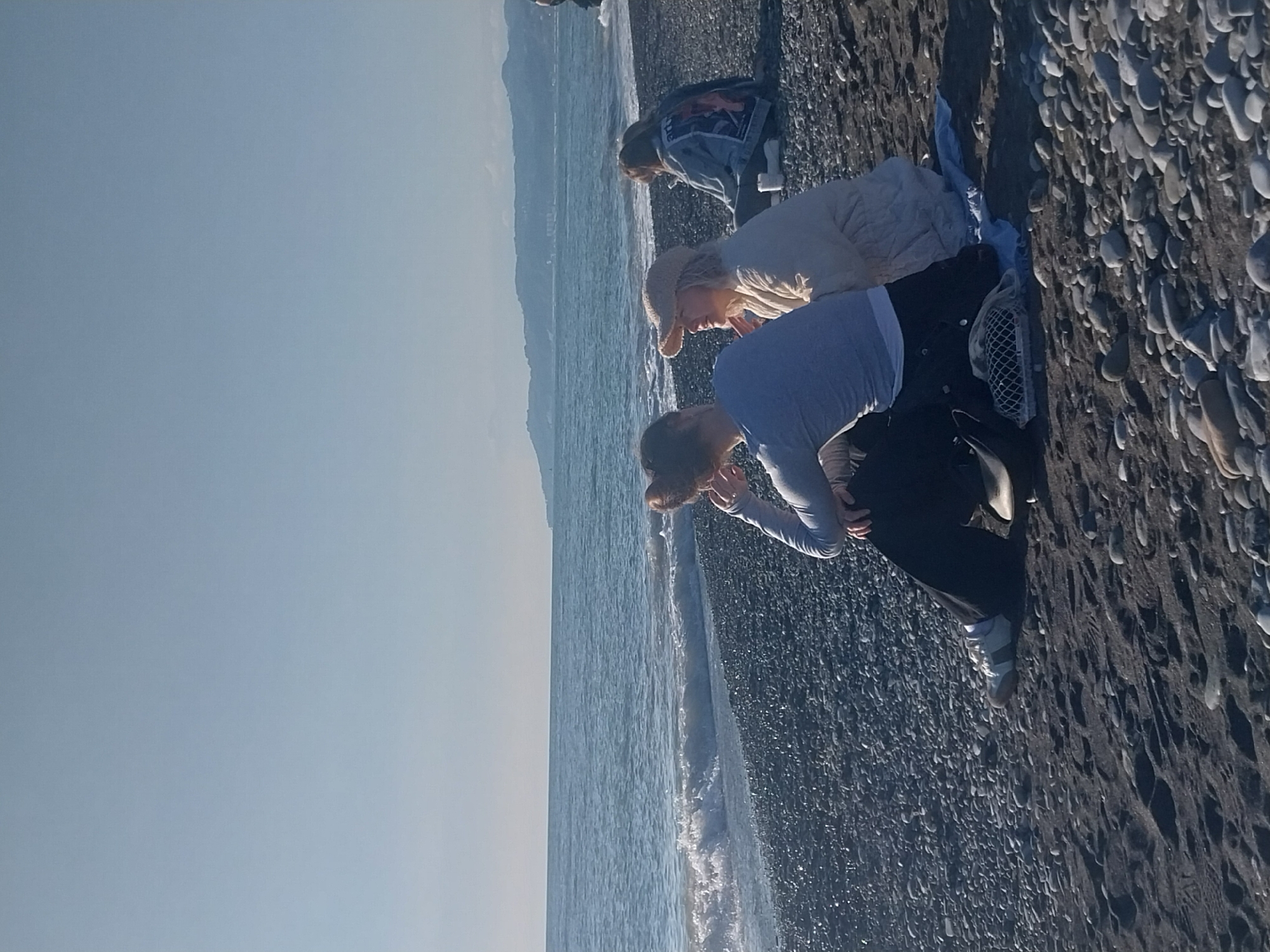 The image captures a sunlit coastal scene in Sochi, Russia, featuring three individuals engaged in an activity on a pebble beach. The sea stretches across the middle ground, bordered by a vast expanse of pebbles on the right and a clear sky to the left, with distant mountains faintly visible across the water.
All three individuals appear to be women, and they are bent over, focusing intently on the ground or the shallow water near the shore. In the foreground, a person with light brown hair, dressed in a light blue long-sleeved top and dark pants, is crouched low. They wear white sneakers, and a mesh bag, likely used for collecting, is visible near their feet on the dark pebbles.
Behind them, a second person, wearing a light beige jacket and a light-colored hat, is also stooped over. A light-colored blanket or piece of fabric is placed on the pebbles next to this individual. Further back and closer to the water, a third person in a denim jacket is similarly bent, peering downwards.
The beach is composed entirely of dark and light gray pebbles. Gentle waves are breaking along the shore, creating a white foamy line. The sky is bright and clear, suggesting a sunny day, possibly late morning or early afternoon. The overall impression is one of calm and focused leisure, perhaps searching for shells, sea glass, or unique stones along the beautiful pebble beach. No visible text is present in the image.