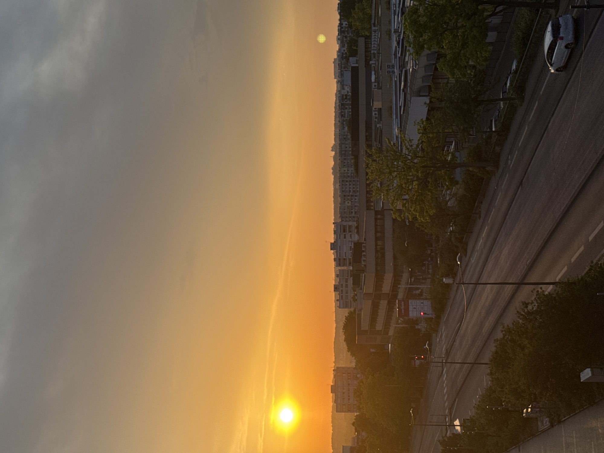 An aerial, long shot captures a city skyline at sunset. The sky is a gradient of warm orange to a pale gray at the top. The sun, a bright yellow orb, is positioned in the upper left corner of the frame. In the mid-ground, several multi-story buildings with flat roofs and uniform windows are visible. Trees with green foliage dot the landscape between and around the buildings. A road runs through the foreground, with lanes marked by white lines. Traffic lights are visible, showing red. A white car is parked on the right side of the road. The scene appears to be an urban environment during the late afternoon or early evening.