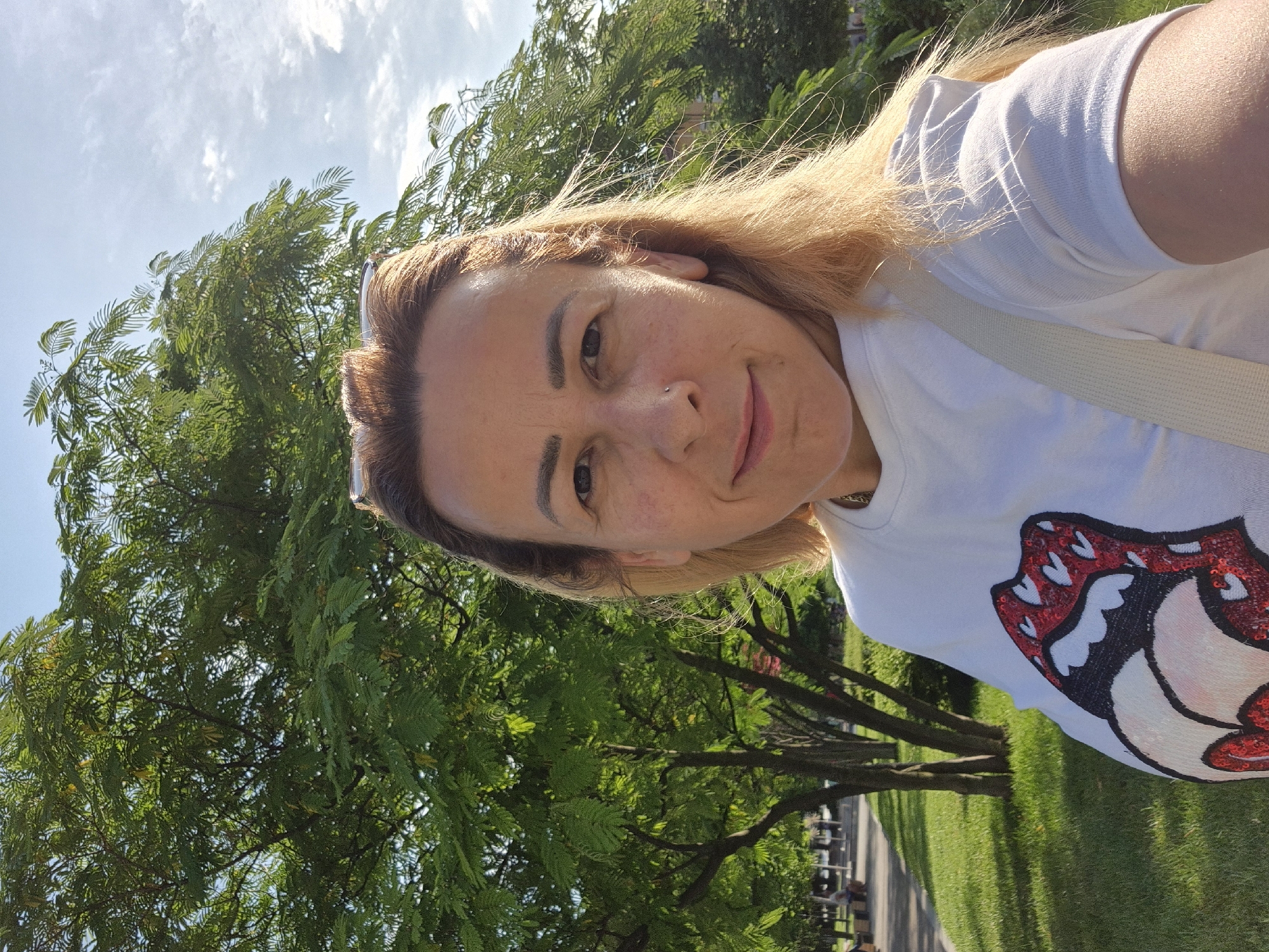 A woman with blonde hair and a white t-shirt with a red and white sequined graphic is standing in front of a tree. She is looking at the camera with a slight smile. The background shows a park with green grass, trees, and a pathway. The sky is blue with some clouds. She is wearing sunglasses on top of her head and has a nose ring.