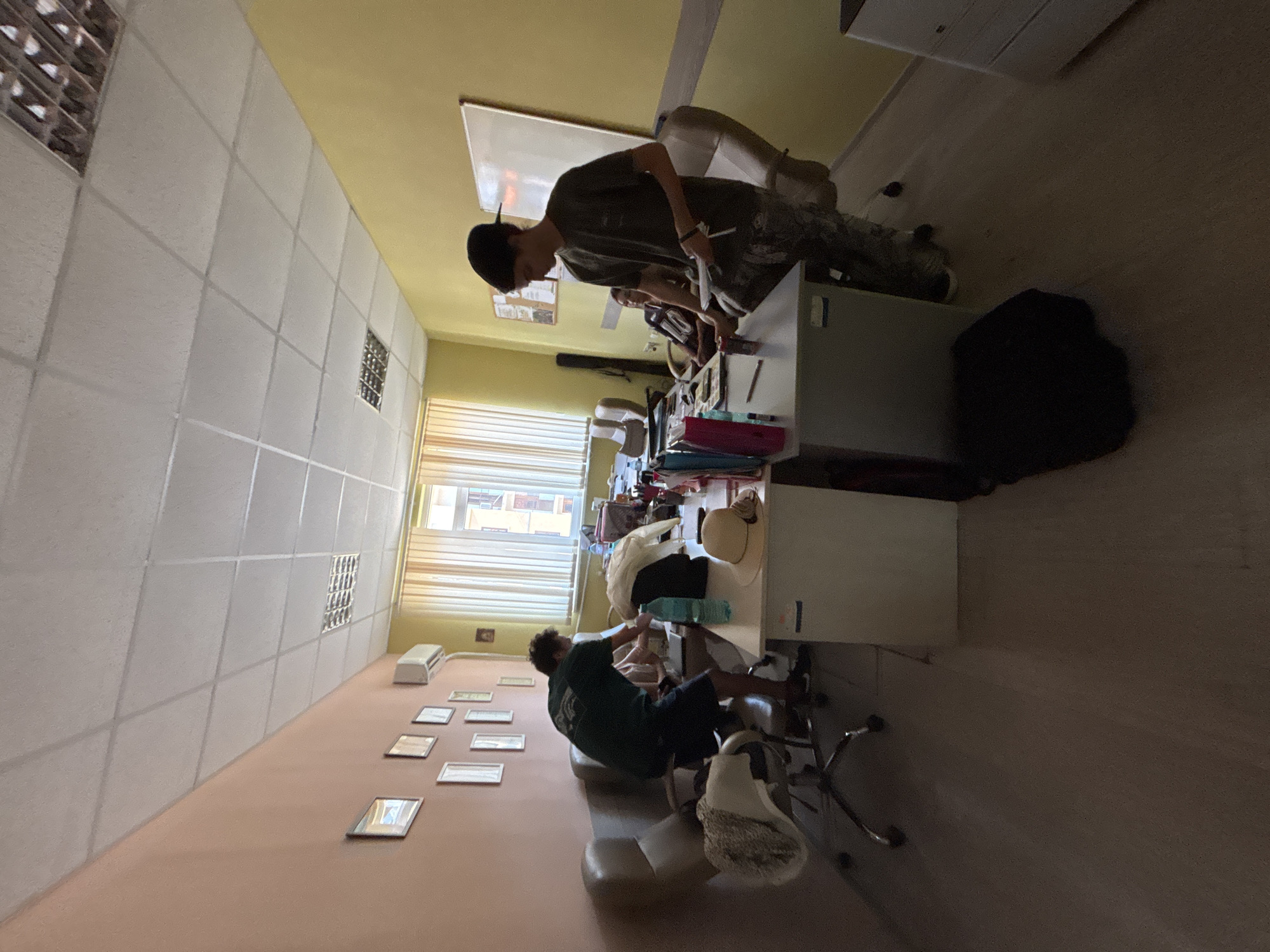 The image depicts an indoor scene, likely an office or classroom, with two young males and one young female visible. The room has pale pink and yellow walls, a suspended ceiling with fluorescent lighting, and wooden blinds on a large window.
On the left, a young male wearing a dark green t-shirt and dark shorts is bending over a desk, seemingly organizing items. He is positioned next to a swivel chair with a patterned cover.
In the center and right, another young male, wearing a dark olive t-shirt and camouflage pants, stands at a desk. He is holding papers and looking towards the right. A young female is seated at a desk behind him, partially obscured.
The desks are cluttered with various objects including bottles of water, folders, papers, and a hat. A whiteboard is mounted on the yellow wall. The floor appears to be linoleum. The environment suggests a casual or educational setting. The image was taken in Vratsa, Bulgaria.