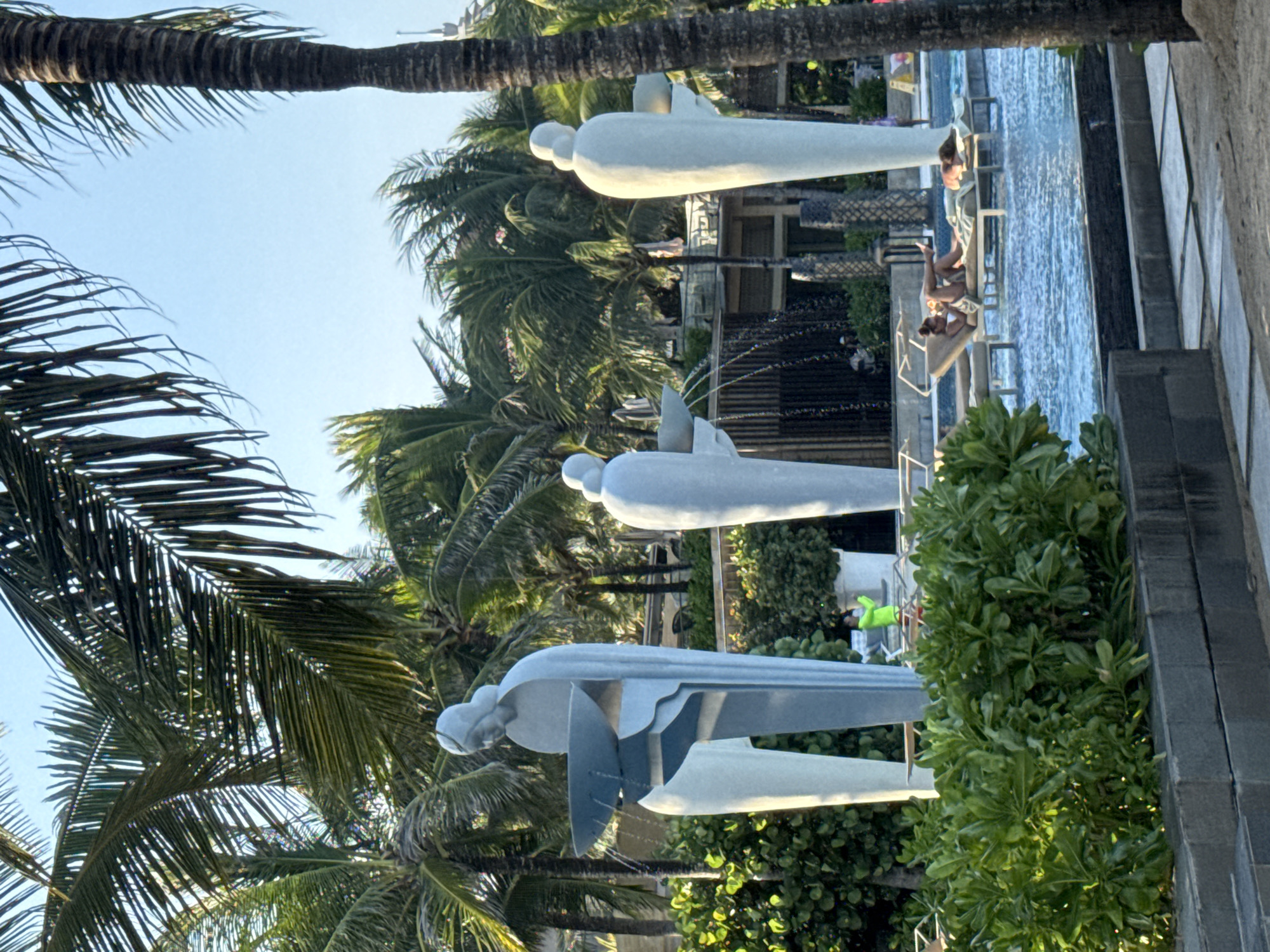 The image depicts a resort or hotel in Kuta Selatan, Indonesia, based on the tropical foliage and architectural style. Three light-colored sculptures, possibly representing stylized human figures or abstract forms, stand near a pool or water feature. Water flows from the sculptures into the pool, where two people are sitting on a ledge. Palm trees frame the scene, adding to the tropical ambience. A person wearing a bright green outfit is also visible near the pool, possibly a staff member or guest.