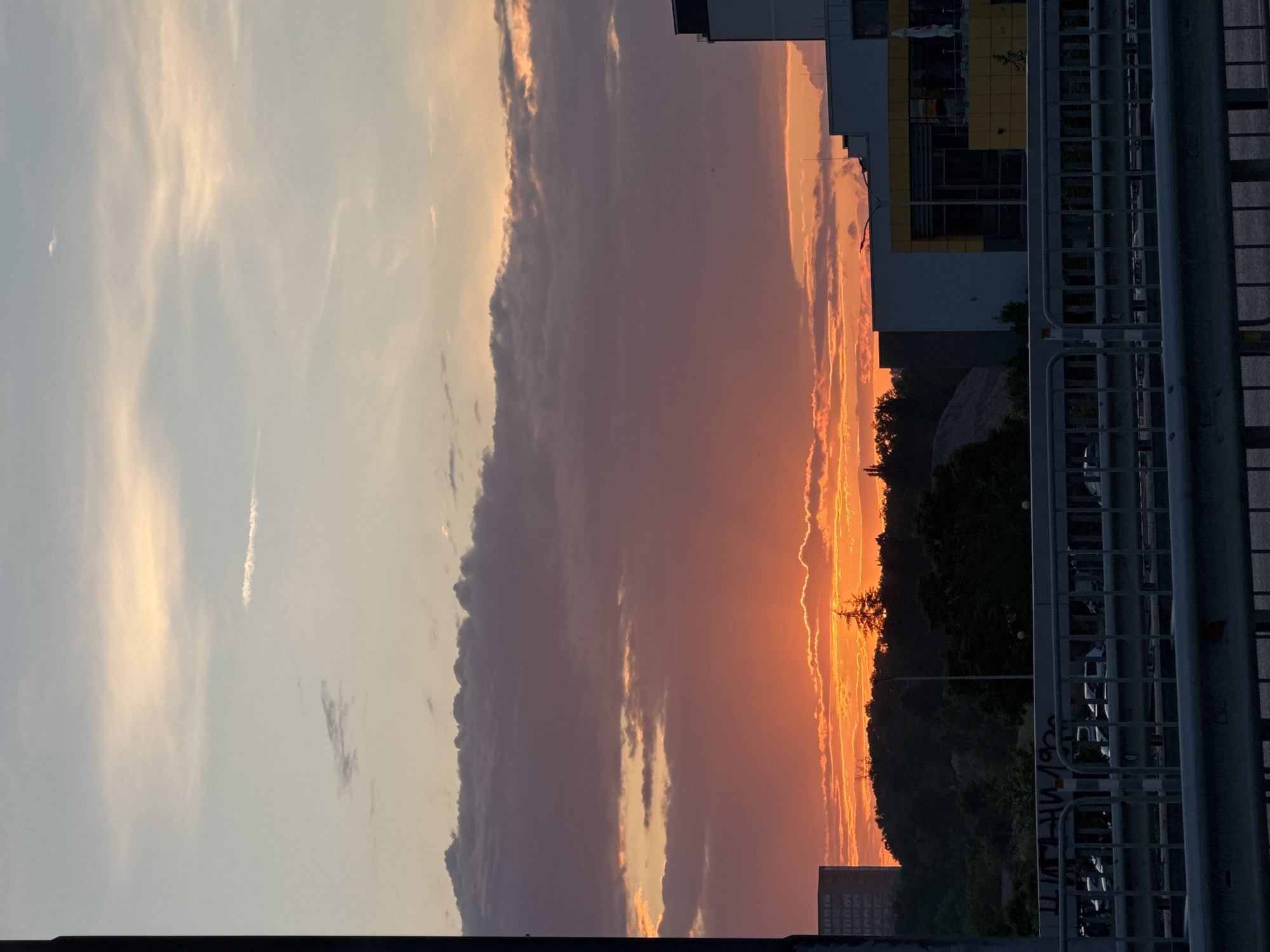 The image displays a dramatic sunset over an urban or suburban landscape in Plovdiv, Bulgaria. The sky is the most prominent feature, showcasing a striking transition of colors from fiery oranges and reds at the horizon, where the sun has just dipped, through deep purples and grays in the middle, to a soft, light blue with delicate white cirrus clouds at the top. A bright, thin contrail is visible in the lighter part of the sky on the left.
A long, dark cloud formation stretches horizontally across the mid-section of the sky, with its lower edge brightly illuminated by the setting sun. Below the vibrant sky, a dark silhouette of a treeline or distant hill is visible, with sparse tree branches sharply outlined against the intense orange glow.
In the mid-ground, several modern buildings with flat roofs and distinct yellow architectural elements are present. The immediate foreground features a robust metal barrier or fence, likely part of a bridge or a roadside structure. Through the gaps in this fence, the tops of parked cars in what appears to be a parking lot are discernible. The scene conveys a serene and majestic atmosphere as daylight fades. No decipherable text is visible.