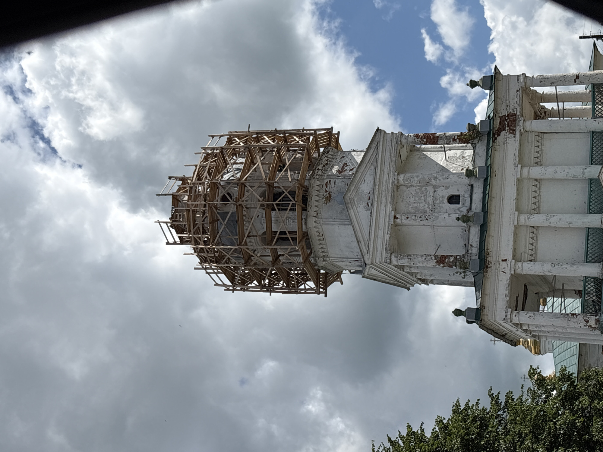 The image shows a close-up view of the upper section of a white church tower undergoing renovation. The tower features classical architectural elements such as columns and pediments, and its white facade is weathered and shows signs of damage, with exposed brickwork in places. A prominent feature is the extensive wooden scaffolding encircling the topmost part of the tower, indicating ongoing construction or repair work. The sky above is partly cloudy, with patches of blue sky visible amongst large, grey clouds. To the left of the tower, a portion of a green tree is visible. The overall scene suggests a historical religious building being preserved or restored. This building is likely the bell tower of the Holy Trinity Church in the village of Kstinino, Kstovsky District, Nizhny Novgorod Oblast, Russia.