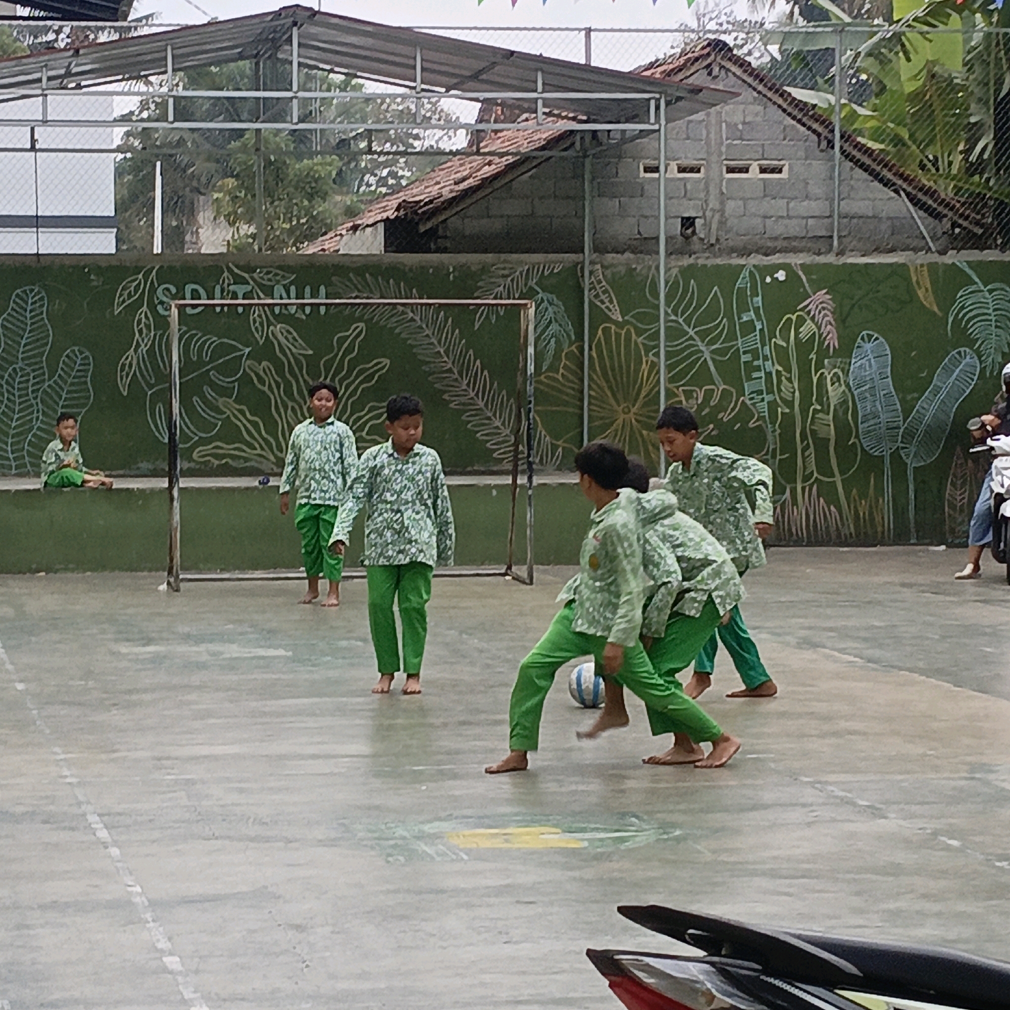A group of young boys, likely students, are playing soccer on a concrete court in front of a green wall decorated with chalk drawings of leaves and vines. The boys are all wearing matching green patterned shirts and pants, and most are barefoot. One boy is sitting on a low green ledge to the left, watching. In the background, a chain-link fence separates the court from trees and buildings. A soccer goal is visible behind the players. Further in the background, a red-tiled roof of a building is visible, and a person on a motorbike is partially visible on the right side of the image. The overall scene suggests an outdoor recreational activity, possibly at a school or community center in Salatiga, Indonesia.