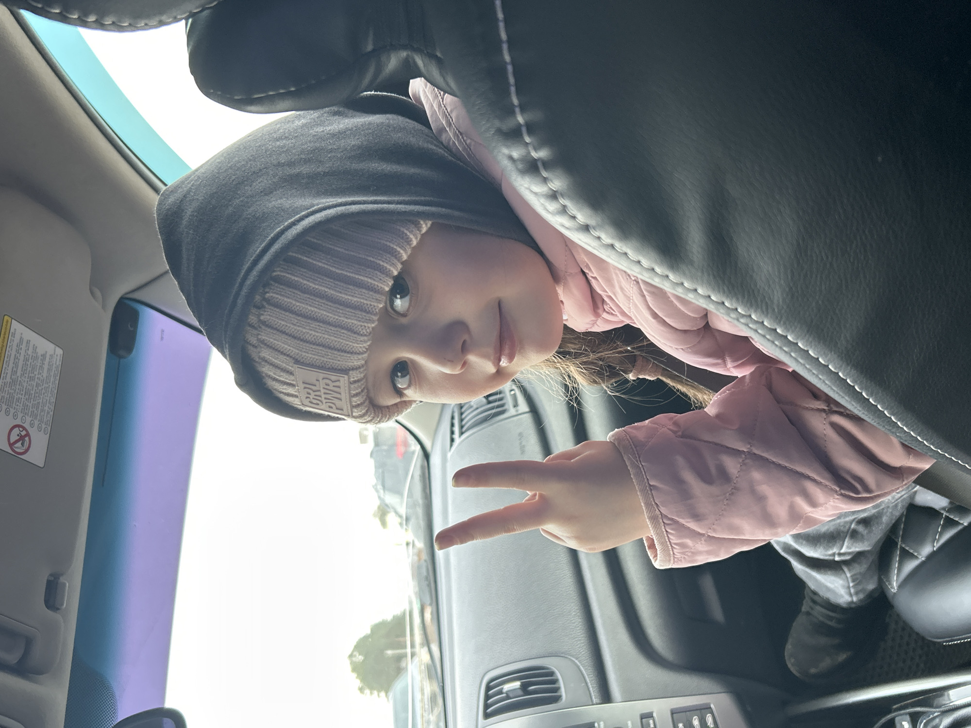 A girl is posing for a selfie inside a car. She is wearing a gray hooded sweatshirt and a pink quilted jacket. On her head, she has a knitted beanie with the words "GRL PWR." She's holding up her fingers in a peace sign. The interior of the car is visible, including the dashboard, seats, and the car's sun visor. Outside the car, the sky is bright, and some trees can be seen in the distance. The exact location within the city of Chaikovsky, Russia is not discernible from the image.