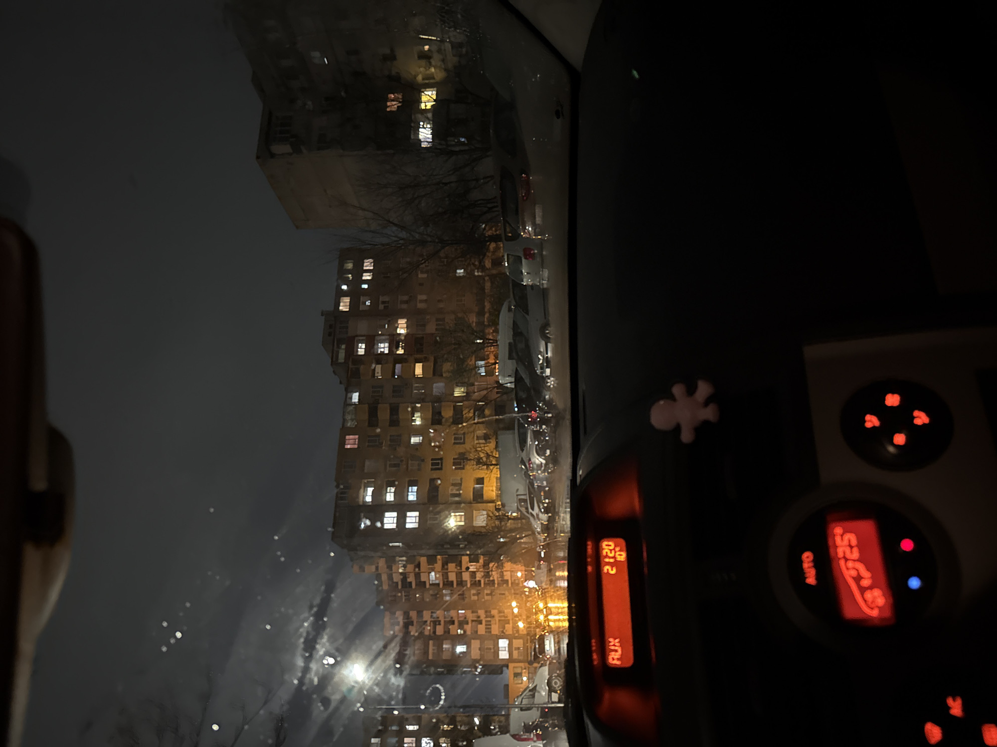 The image captures a nighttime urban scene from within a vehicle in Plovdiv, Bulgaria. The perspective is through a wet front windshield, displaying numerous water droplets that refract light sources. The vehicle's dashboard occupies the lower portion of the frame. A central digital display unit on the dashboard shows "AUX," "21:20" (indicating the time), and "10°" (indicating the external temperature). Directly beneath this, a climate control panel with illuminated red buttons and controls displays "AUTO" and a set interior temperature of "25°." A small, light pink, humanoid-shaped figurine is positioned on the right side of the dashboard.
Beyond the vehicle, multiple multi-story residential apartment buildings, characteristic of Eastern European architecture, dominate the background. Many windows across these structures are illuminated. A street or parking area in front of the buildings contains numerous parked vehicles. Artificial light sources, including streetlights, cast distinct orange and yellow reflections on the wet ground surface. Bare trees are interspersed between the buildings and the parked vehicles. No individuals are visibly present in the exterior scene.