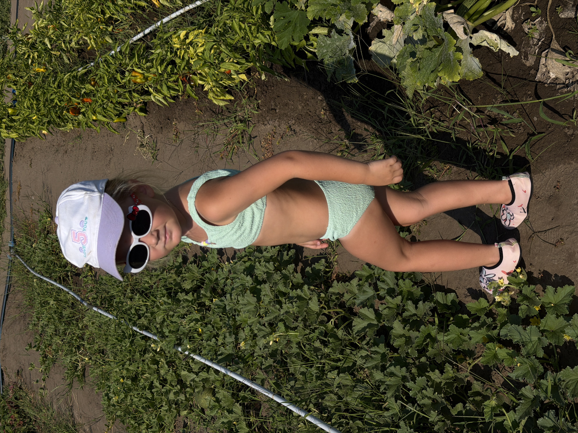 A young girl wearing a light green bikini, a white and purple baseball cap, and pink water shoes with floral patterns stands in a garden. She has on large, round, black sunglasses and red Mickey Mouse-shaped earrings. The girl is looking towards the camera with a slight smile. The garden appears to be growing peppers and squash. A white hose runs along the ground in front of her. The scene suggests it is a sunny day. The image appears to be taken in Kamyshin, Russia. The baseball cap has the number "5" and the text "Love you" printed on it.