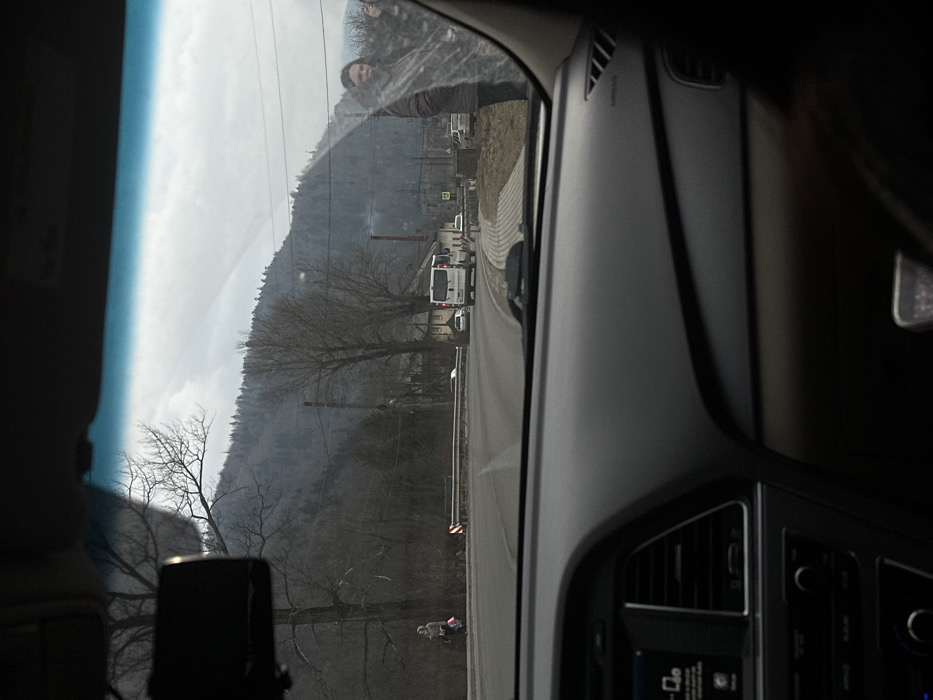 The image depicts a view from the interior of a vehicle, looking forward through the windshield onto a road in Skole, Ukraine, under an overcast sky. The vehicle's dark gray dashboard, central infotainment screen, and air vents are visible in the foreground, along with a rearview mirror mounted at the top center. The windshield exhibits smudges and reflections, particularly prominent on the right side.
A paved, two-lane road curves to the right, extending into the midground. A metal guardrail follows the left edge of the road, while a sidewalk constructed of light-colored pavers is visible on the right. Utility poles with overhead wires line both sides of the road. A white and red chevron sign indicates the curve on the left, and a white square sign with a black pedestrian icon is positioned near the sidewalk on the right.
On the far left, a person wearing a light-colored coat and head covering walks away from the camera along the road's shoulder, carrying multiple bags. On the right, partially obscured by the windshield's imperfections, a person with dark hair and a dark jacket stands on the paved sidewalk, looking towards the camera with a slight smile. A white cargo van is observed traveling away from the camera in the right lane, followed by a partially obscured second vehicle. Several other vehicles are visible further down the road.
The background features bare deciduous trees along the road, transitioning into a dense, dark forest covering steep hills or mountains. A cloudy sky dominates the upper portion of the frame. Some buildings and a faint plume of smoke are discernible among the trees in the distant midground.