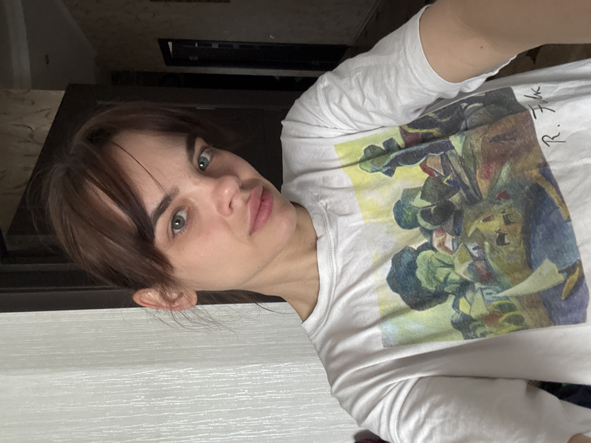 A portrait of a woman with fair skin, green eyes, and brown hair with bangs. She's wearing a white t-shirt with a print that includes the signature "R. Fulk". The background includes a light-colored wall and a dark-toned object or piece of furniture above her head. The photograph's perspective is slightly angled. The location information is not evident in the image.