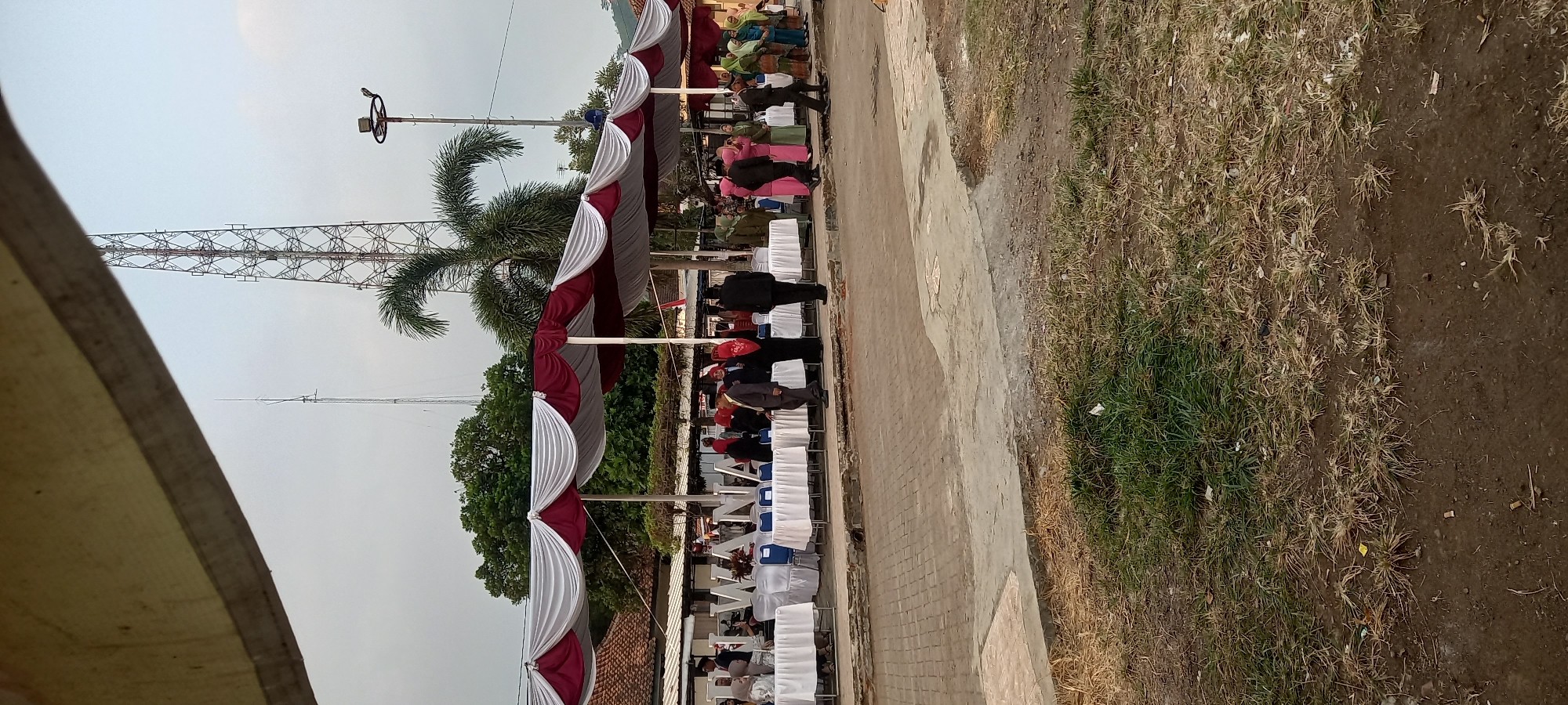 In an outdoor setting at a location possibly related to the sign visible which reads "WANA," likely indicating Wanareja, Indonesia, a gathering is taking place under a red and white striped canopy. Rows of white-covered tables are set up, with some people, dressed in formal attire including headscarves and suits, mingling around them. In the background, a large transmission tower stands tall against a pale sky, with a palm tree in the foreground. The ground is a mix of paved brick and dry grass. The scene appears to be an event or ceremony.