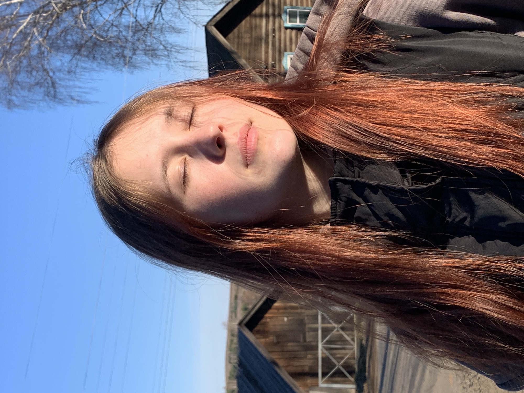 A young woman with long auburn hair is posing outdoors with her eyes closed, likely in bright sunlight. She is wearing a dark-colored jacket with a gray hoodie underneath. Behind her, there are old wooden buildings, potentially barns or sheds, typical of rural areas. The background also includes a clear blue sky with some overhead power lines visible. The location appears to be in or near the city of Фоки, Russia, based on the presence of structures that might be found in the region.