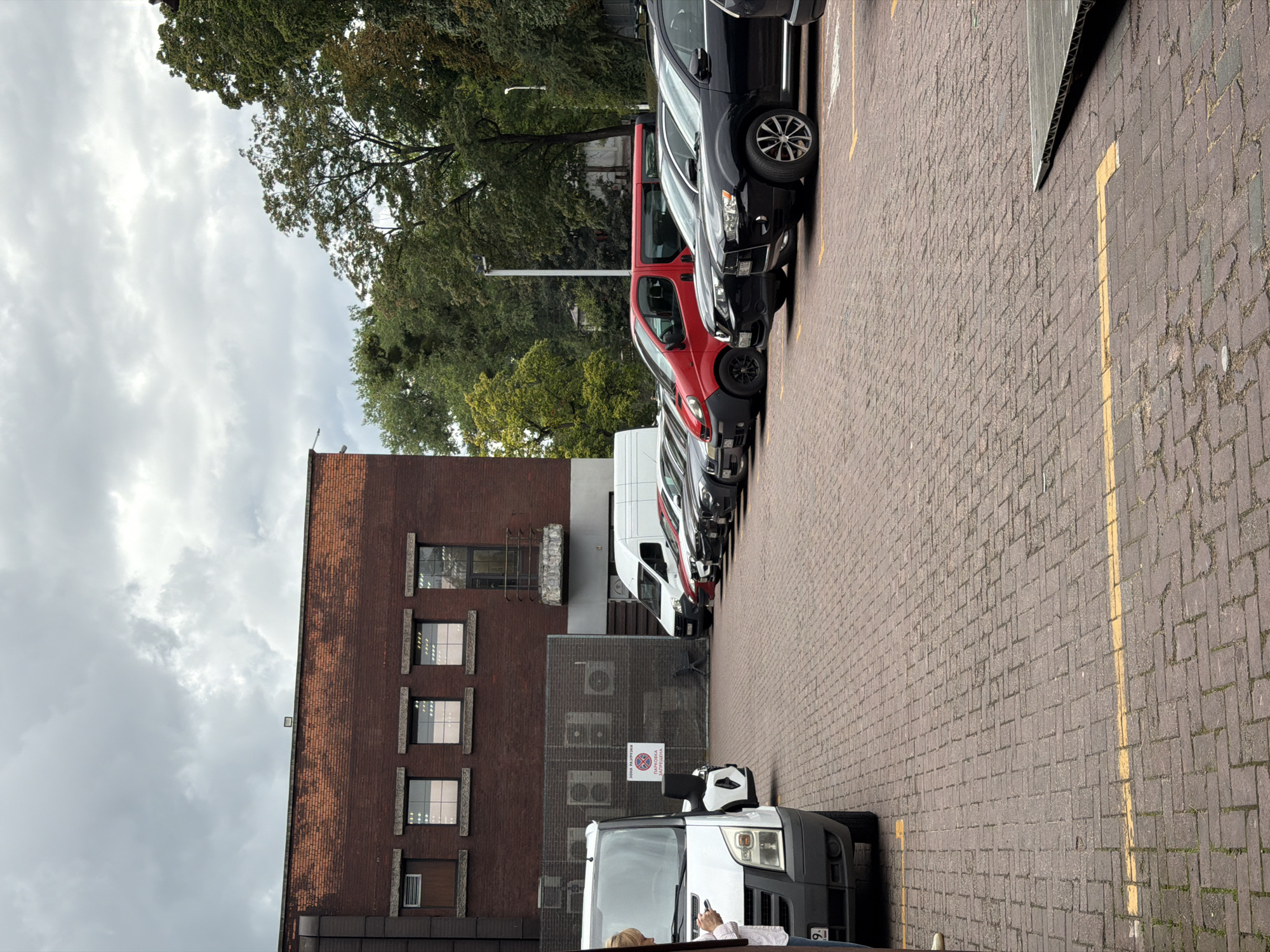 The image depicts a parking lot with a brick-paved surface, marked with yellow lines. A brick building with multiple windows and an air conditioning unit is visible in the background. Several vehicles are parked in the lot, including vans and cars of various colors. A person is standing next to a white van in the foreground, looking at a mobile device. The sky is overcast with clouds. The image does not contain any elements that would suggest it was taken in Kaliningrad, Russia.