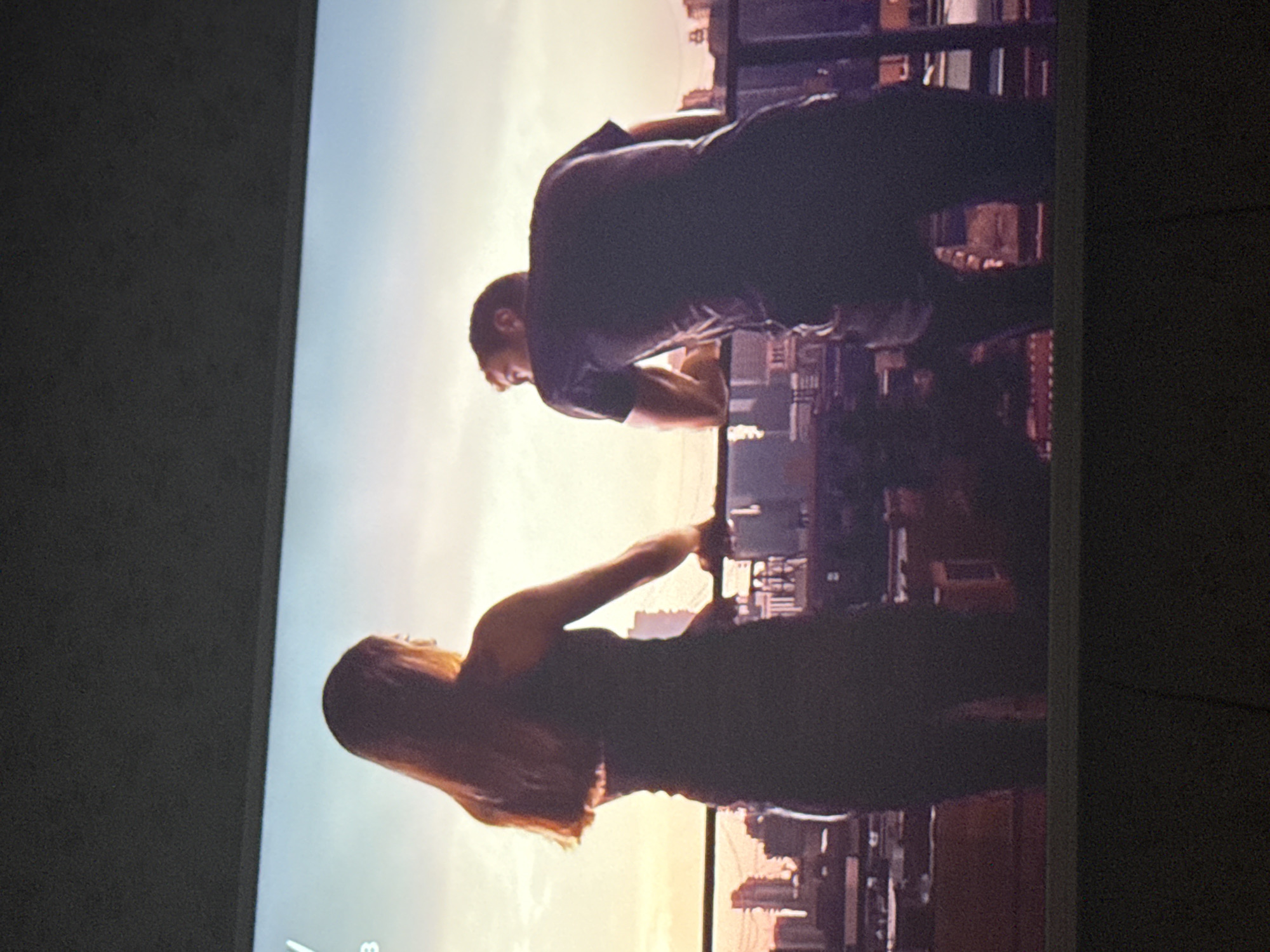 A man and woman are standing on a balcony overlooking a cityscape. The man is leaning forward with his hands on the railing, while the woman is standing behind him, reaching out to touch his hand. The background shows buildings at sunset, with a color transition from blue to orange. There is no indication this picture was taken in Mytishchi, Russia.