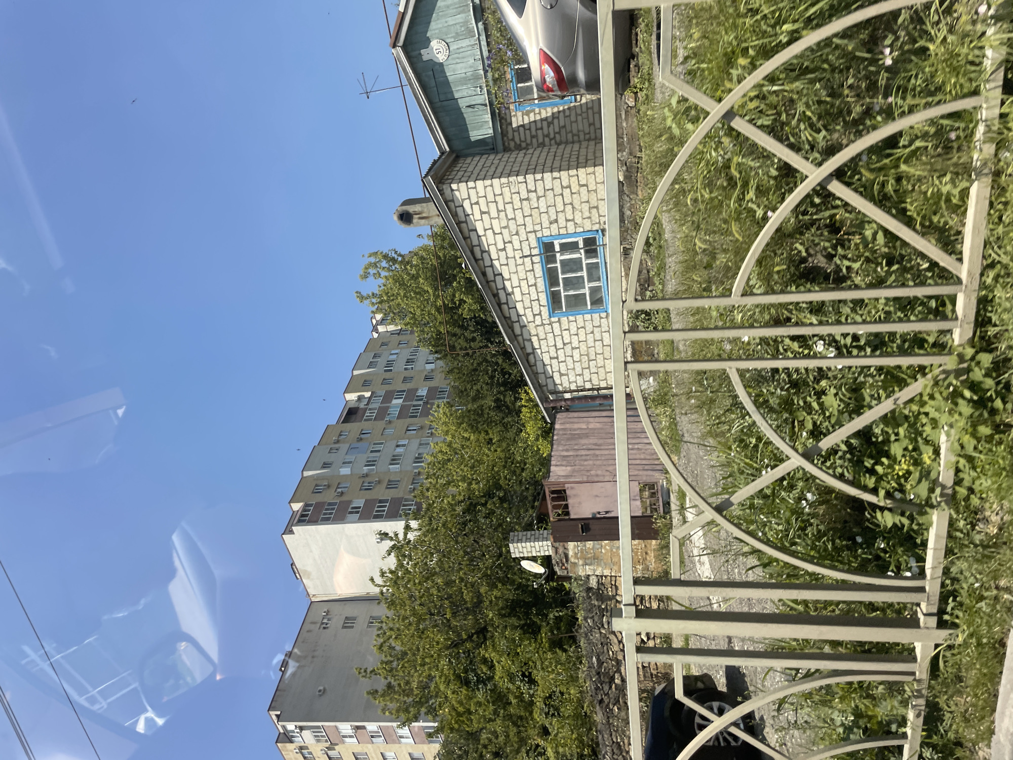 The image shows a residential area in Stavropol, Russia. In the background, a multi-story apartment building is visible, constructed with light-colored facades and multiple rows of windows. In front of the apartment building, there are dense green trees. Closer to the foreground, a single-story house made of white brick with a green roof is situated. This house has blue-trimmed windows and a visible chimney. A silver car is parked in front of the house. A metal fence with decorative arches runs across the lower part of the image, obscuring the immediate foreground which is filled with green grass and some small white flowers. The sky is clear and blue. The image appears to be taken from a moving vehicle, as indicated by the reflections on a glass surface in the upper portion of the image.