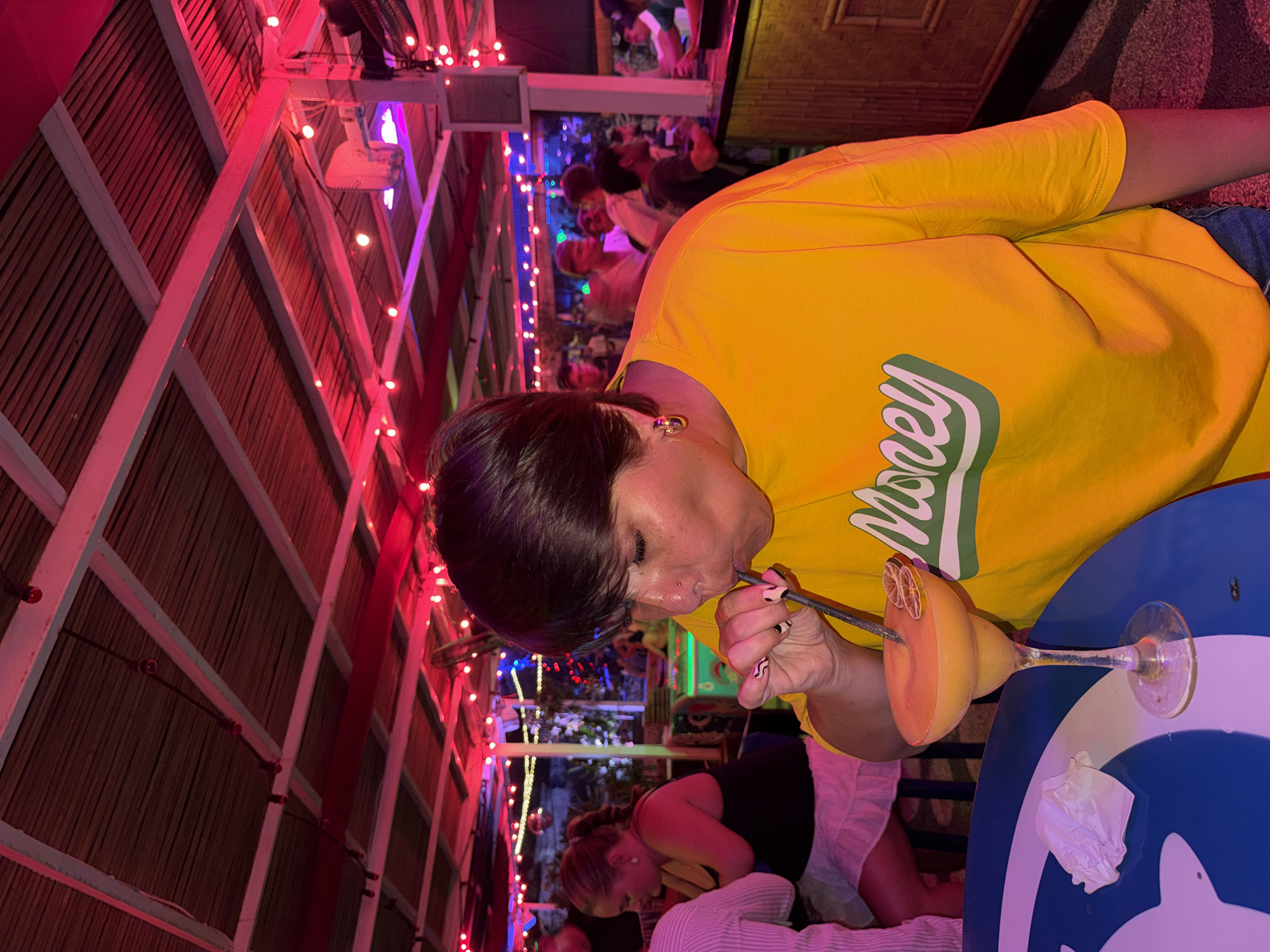A woman in a yellow "Money" shirt sips a yellow drink with a straw at a table at a bar. Another woman sits nearby in a black tank top and white skirt. Behind them, other patrons are visible in the crowded, indoor/outdoor bar. The bar has a bamboo-lined roof structure crisscrossed with red and pink string lights. This scene is located in Canggu, Indonesia.
