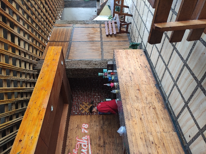 The image shows a wood-paneled counter in a cafe or restaurant, with a worker in red preparing beverages. The counter has a dark countertop and a wooden facade with a sign that says "udh 78". There is a brick wall in the background. The cafe has a tiled floor and a bamboo-covered ceiling. To the right of the counter, there are two tables and chairs outside. There is a wooden bench in the foreground of the image, and there is a planter with greenery next to the counter. The scene takes place at latitude -7.855 and longitude 112.454, which is in Kecamatan Pujon, Indonesia.