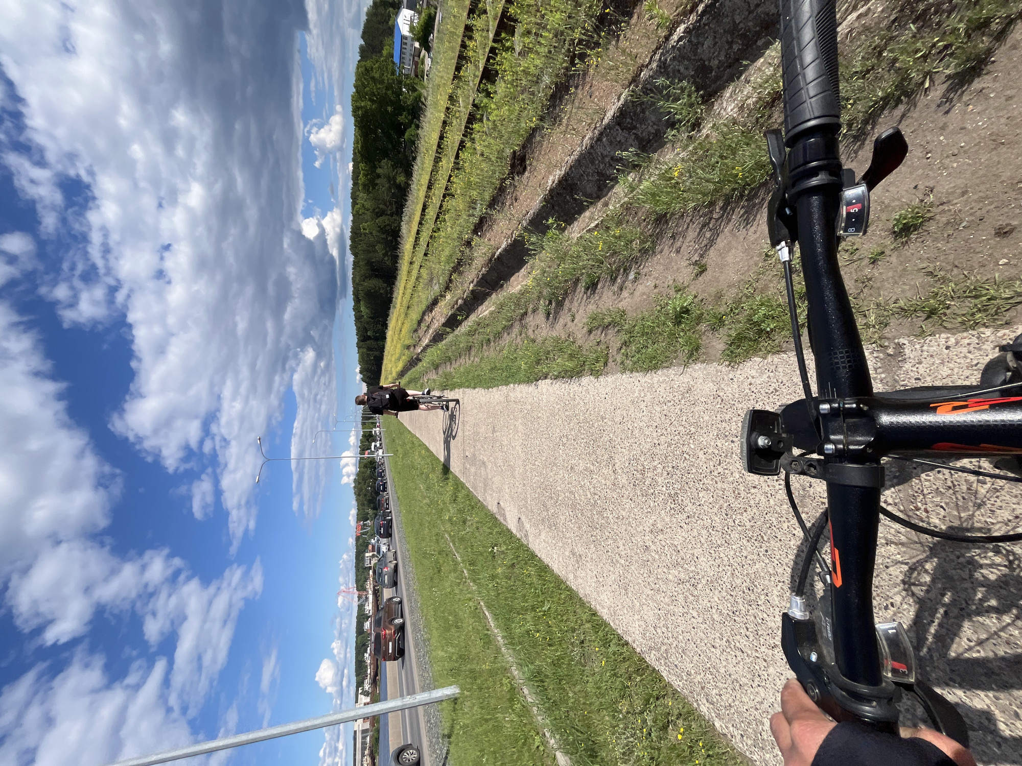 The image shows a first-person perspective of someone riding a bicycle on a paved path. To the left of the path is a busy road with cars in traffic. To the right of the path are abandoned railway tracks overgrown with grass and weeds. In the distance, a line of trees forms the horizon. The sky is blue with scattered white and gray clouds. A lone cyclist can be seen further down the path in the distance. The location is Chaikovsky, Russia, given the context of the provided crops hinting at specific landmarks and vegetation.