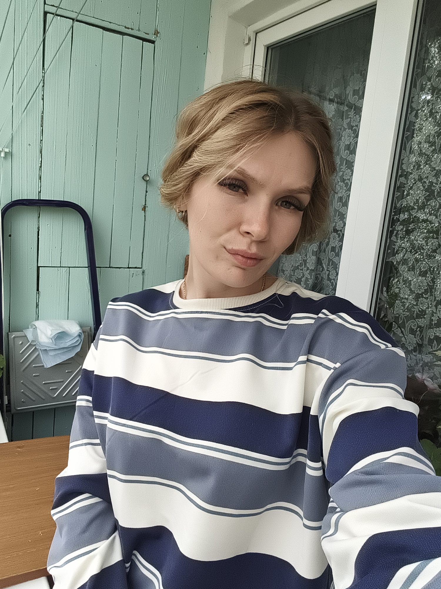 A young woman with blonde, bobbed hair is taking a selfie outdoors. She is wearing a long-sleeved, horizontally striped sweater in shades of navy blue, light blue, grey, and white. Her hair is styled casually, and she has makeup on, including mascara and lipstick. She is standing in front of a pale green wooden wall and a window with a patterned curtain. To her left, a blue ladder is leaning against the wall, with a tray holding folded white cloths beneath it. A portion of a wooden surface is visible in the lower left of the frame. The setting appears to be a residential area in Izhevsk, Russia.