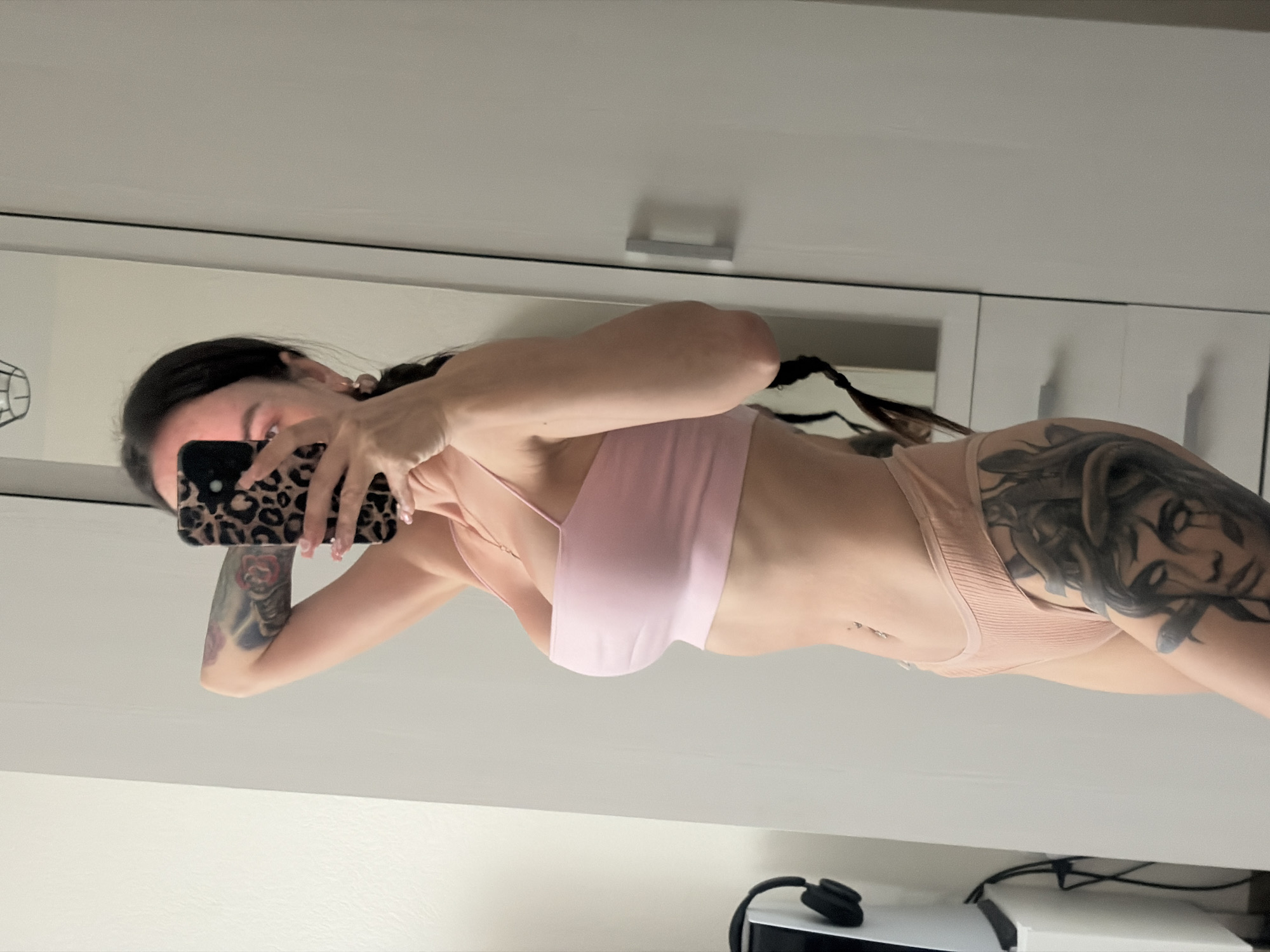A woman is standing in a room, taking a mirror selfie. She has dark hair, braided and hanging down her back. She is wearing a pink crop top and nude-colored underwear. She has tattoos on her left arm and right thigh. In the background, there is a white wardrobe and a mirror reflecting the woman. To the left of the frame, a white stand with black headphones is visible. The room appears to be a bedroom or dressing area.
