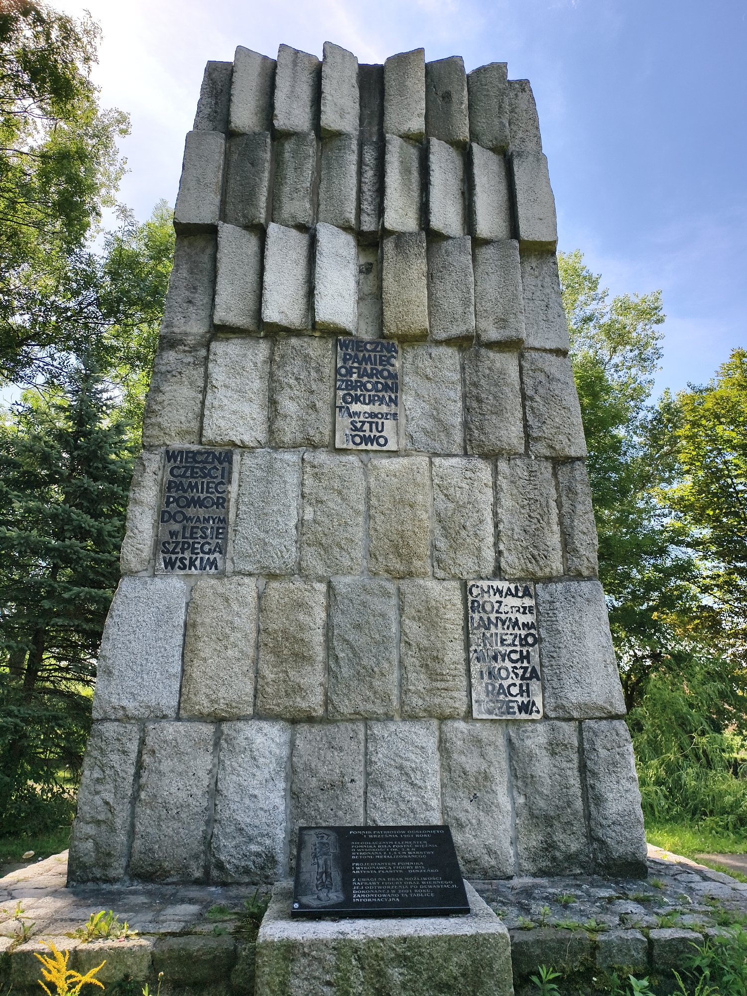 The image shows a tall, monolithic stone monument located in Tczew, Poland. The monument is constructed from large, rectangular stone blocks arranged vertically, creating a textured, stepped appearance at the top. Inscriptions in Polish are carved into some of the stone blocks. On the front of the monument, towards the bottom, there is a dark plaque with further text. The monument is situated outdoors, surrounded by lush green trees and foliage under a clear blue sky. The ground around the base of the monument is covered in grass and small plants, with some loose stones. The overall scene suggests a memorial or historical site in a natural setting.