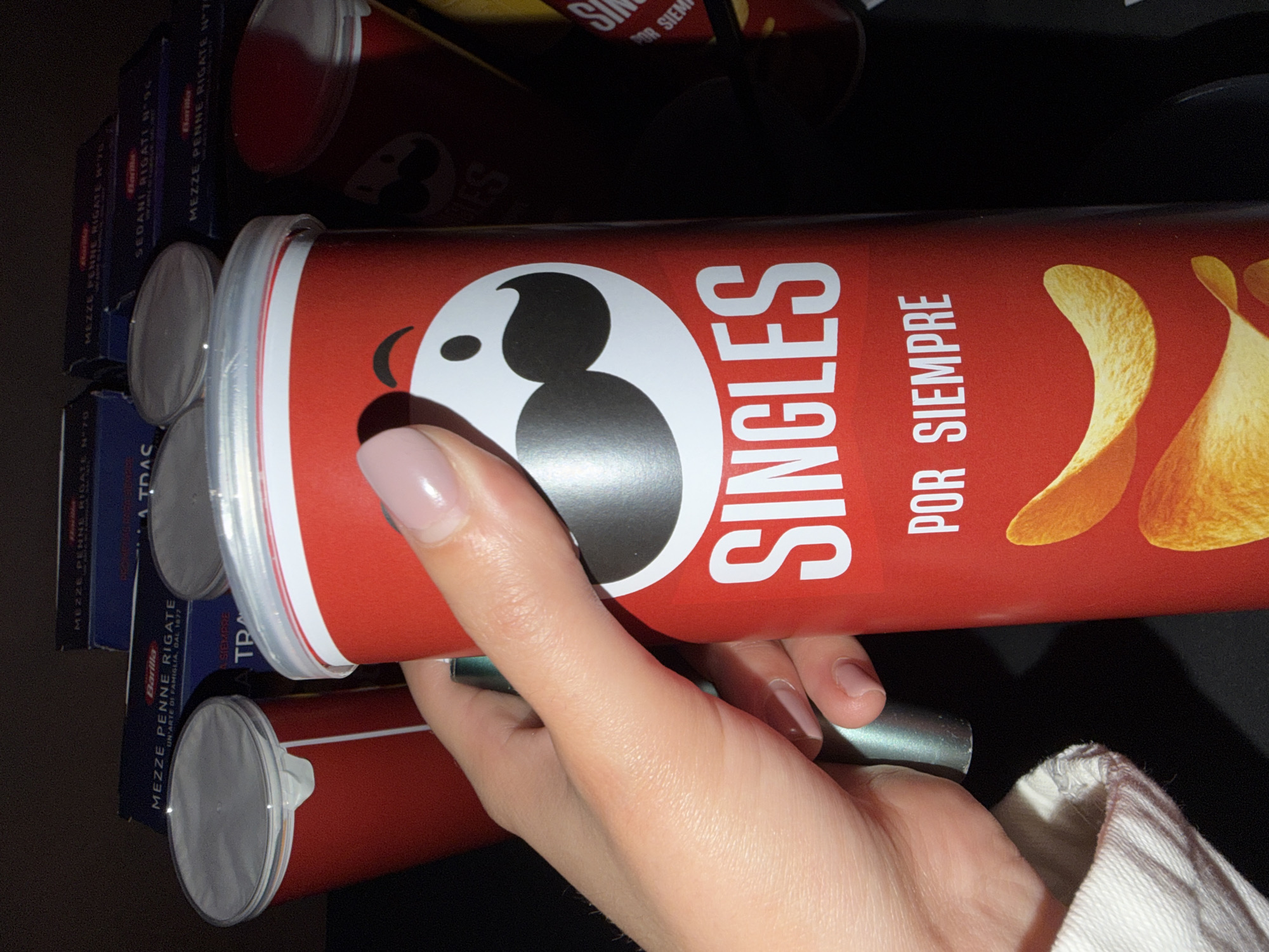 The image shows a hand holding a can of Pringles Singles crisps. The can is red and features the Pringles logo and the words "SINGLES POR SIEMPRE" in white. The hand has a neutral-colored nail polish. In the background, partially visible, are stacked boxes of Barilla Mezze Penne Rigate pasta. The scene appears to be indoors and has a dark background. There is no indication of it being in Milan or any other specific location.