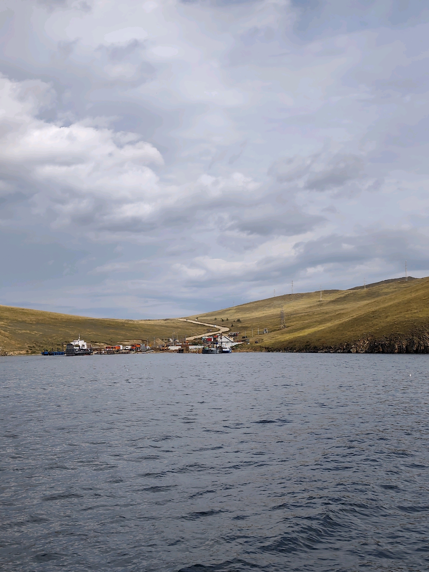 The image depicts a waterfront scene in Khuzhir, Russia, under a cloudy sky. A body of water, likely Lake Baikal, fills the foreground with gentle ripples. Along the shoreline, a small settlement is visible, featuring several boats docked at a pier. Structures include a ferry, a white boat, and various other vessels. Behind the waterfront, rolling hills covered in dry, golden-brown grass stretch upwards. A road winds its way up the largest hill, with several utility poles scattered across the landscape. The overall impression is one of a remote, rugged coastal environment.