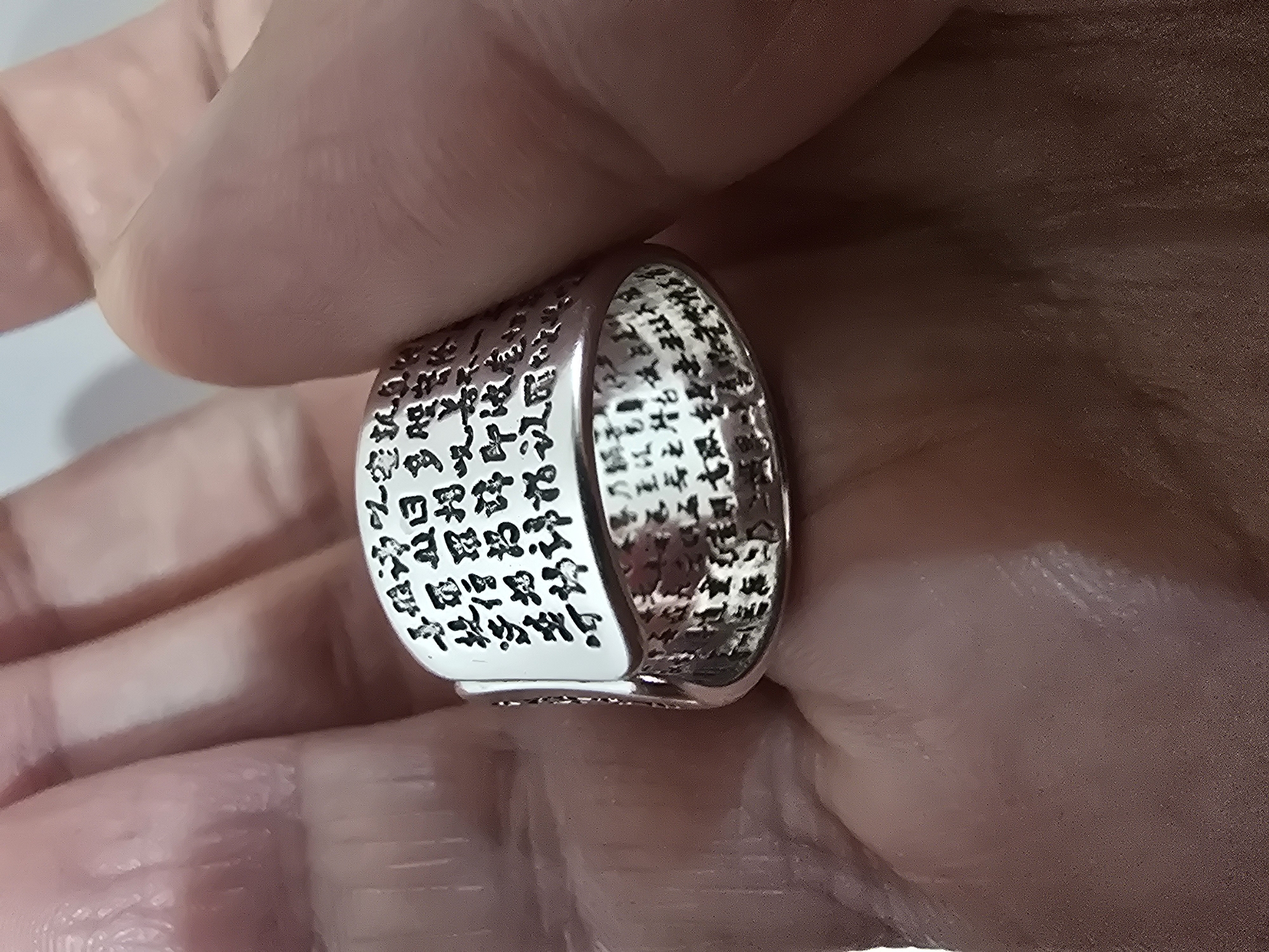 A silver ring with intricate Chinese calligraphy is held in a person's palm, with portions of another hand visible. The ring is positioned on the side, with the inscriptions facing outward and upward. The calligraphy appears to be in black ink and covers the entirety of the ring's surface, both on the exterior and interior. The background consists of the skin of the hand holding the ring. There is no discernible location in the city of Ulan-Ude, Russia, present in this image.