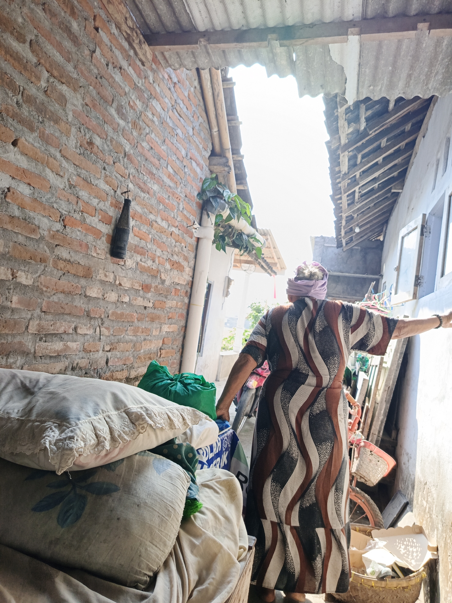 The image depicts an outdoor alleyway in Rowokangkung, Indonesia, characterized by a dilapidated corrugated metal roof and a brick wall. In the foreground, a stack of pillows and fabric items are visible. A woman wearing a patterned dress and a headscarf stands in the mid-ground, facing away from the camera and towards the right side of the alley. Her right arm is extended, touching the wall. Behind her, various objects are present, including a bicycle with a basket, laundry, and storage containers. The scene suggests a residential or storage area with signs of wear and tear, illuminated by bright daylight filtering through the gaps in the roof.