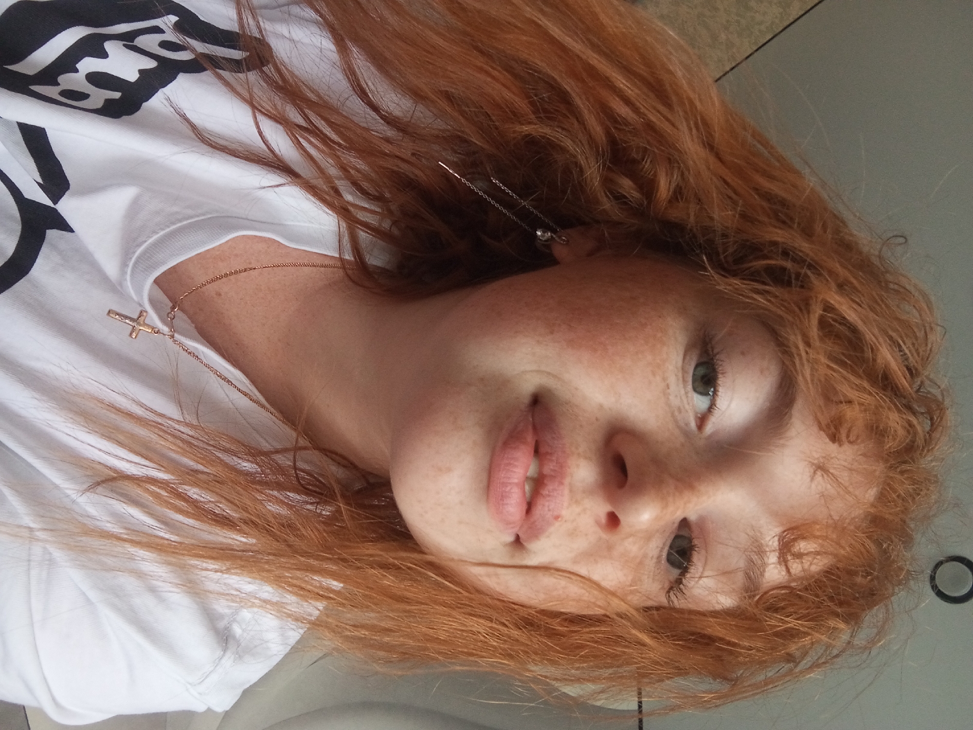The image is a close-up of a young woman with red hair and freckles, wearing a white t-shirt with black print. She is also wearing a gold necklace with a cross pendant and silver chain earrings. The background appears to be a smooth surface, possibly a table or floor. The photo's location is Unknown, Unknown.