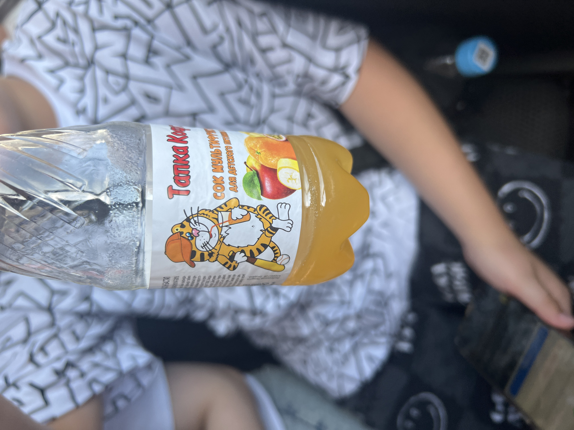 The image shows a close-up of a plastic bottle filled with yellow liquid. The bottle has a label with a cartoon tiger wearing an orange cap and holding a straw. The label also depicts images of an apple, an orange, and a banana, and text in Russian that reads "Татка Коф" and "СОК МУЛЬТИФРУКТ". The bottle is held by a child whose arm and shoulder are visible on the right side of the frame. The child is wearing a white t-shirt with a black pattern. The background is blurred but appears to be an indoor setting, possibly a car. The overall lighting suggests it is daytime. There is no specific information in the image that would indicate its location in the city of Ulyanovsk, Russia.