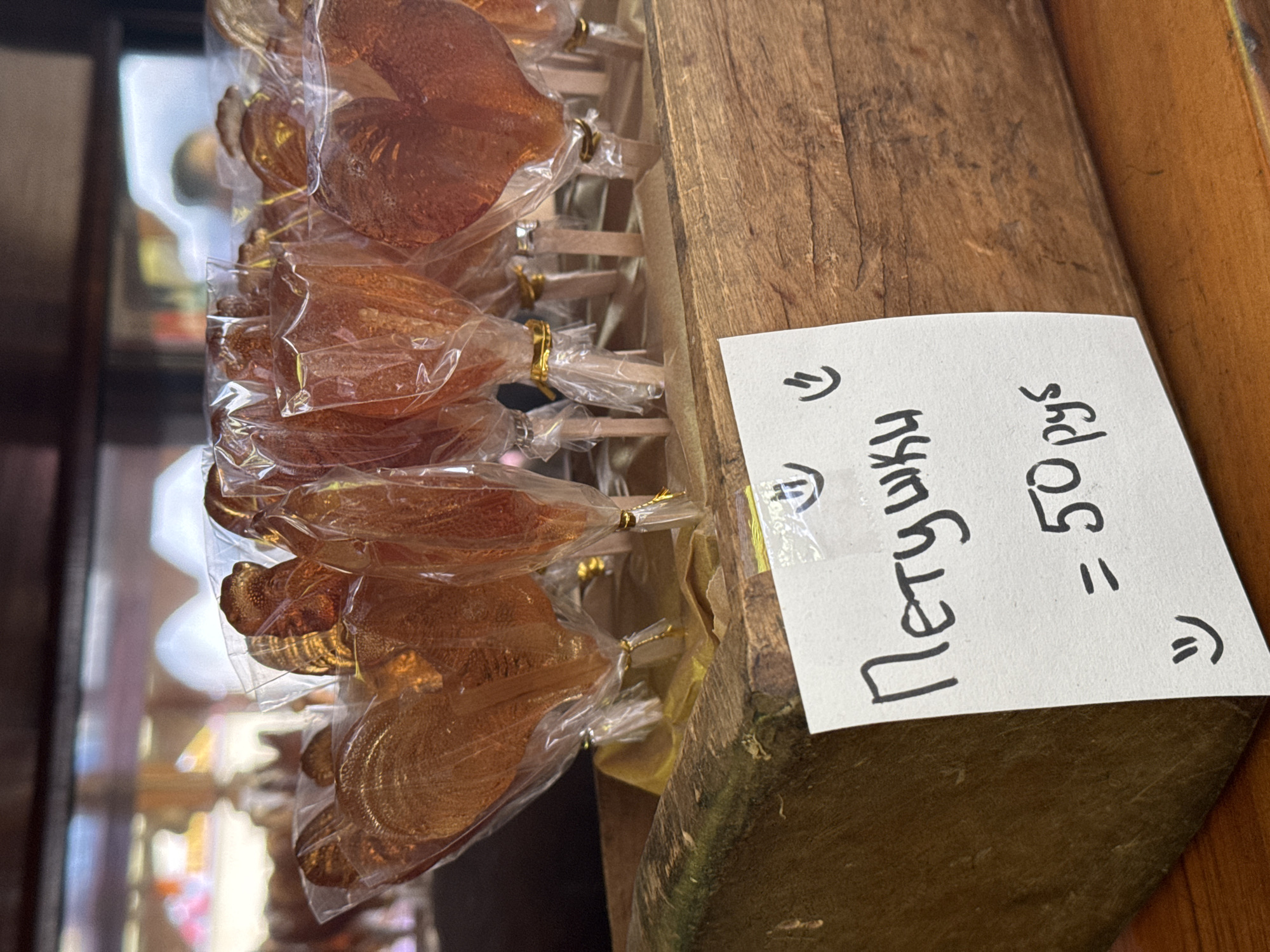 The image shows a display of lollipops in a wooden container with a sign in Russian. The sign reads "Петушки = 50 руб," meaning "Lollipops = 50 rubles." The lollipops are packaged in clear plastic and appear to be shaped like cockerels. The location is not discernible in the image.