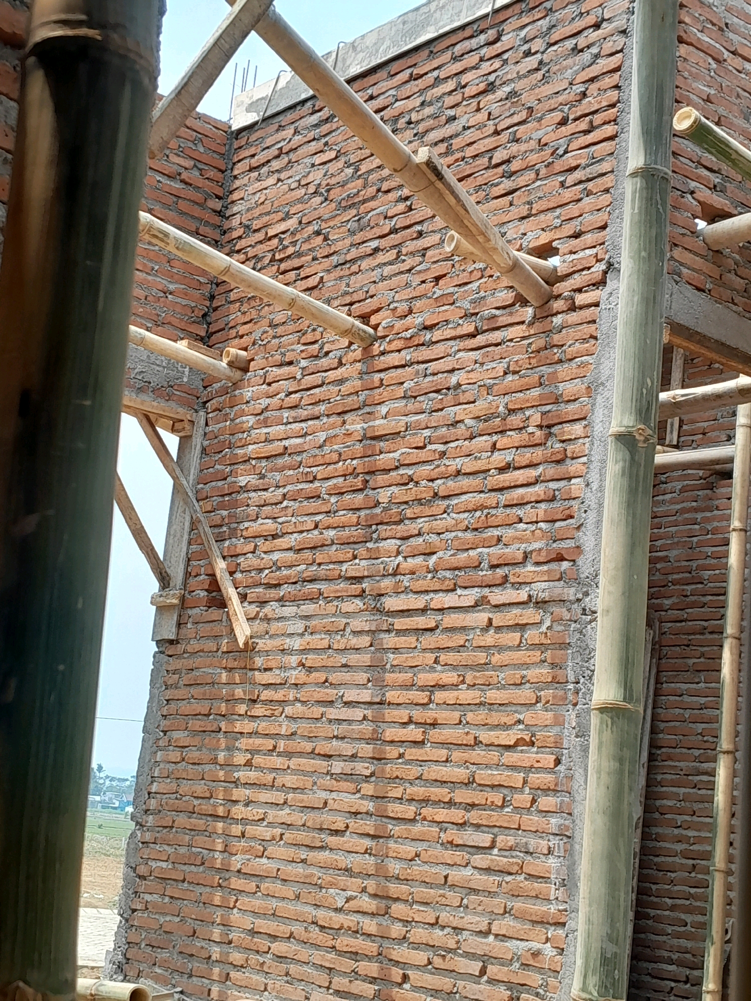 The image shows a brick wall under construction with bamboo scaffolding. The wall is made of red bricks and mortar. The bamboo poles are positioned vertically and horizontally, supporting the construction. In the background, there is a glimpse of a rural landscape with some buildings and greenery, suggesting the location is Karangduren, Indonesia. The sky is visible above the construction. No people are present in the frame.