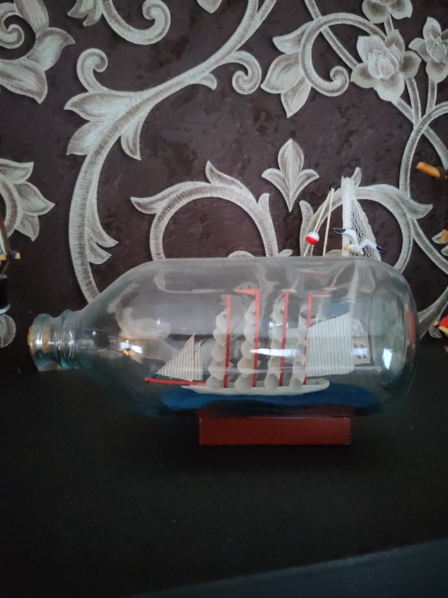 A ship in a bottle sits on a dark surface, possibly a shelf or table, with a patterned wallpaper in the background. The bottle is made of clear glass and lies horizontally, revealing a miniature sailing ship inside. The ship has white sails with red masts and a blue hull. Two small model seagulls are perched on top of the bottle, near the neck, along with a red and white buoy. The wallpaper behind the bottle features an ornate, light-colored floral or scroll pattern against a darker, textured background. The scene is well-lit, highlighting the details of the ship and its enclosure. No people are visible in the image. There is no information available about the location in the city of Bol'shie Mkhi, Russia.