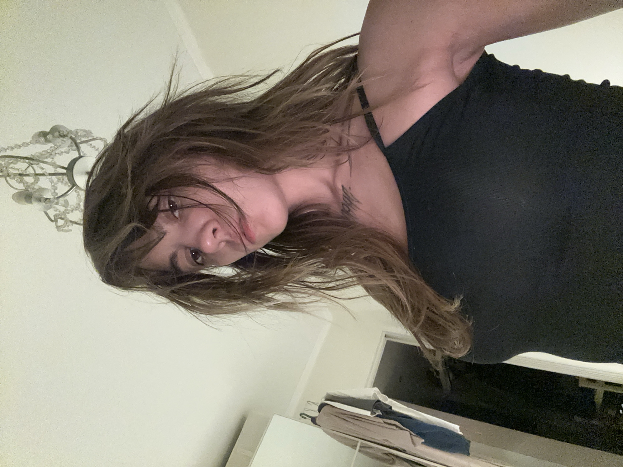 The image shows a young woman with long, messy brown hair and bangs, looking down at the camera. She is wearing a black tank top. Her left shoulder is visible. In the background, a chandelier hangs from the ceiling. To the left of the woman, there is a closet with clothes hanging on the door. A doorway is visible in the distance. The scene appears to be indoors, in what could be a bedroom or a dressing area. There are no indications in the image to suggest it is located in Moscow, Russia.
