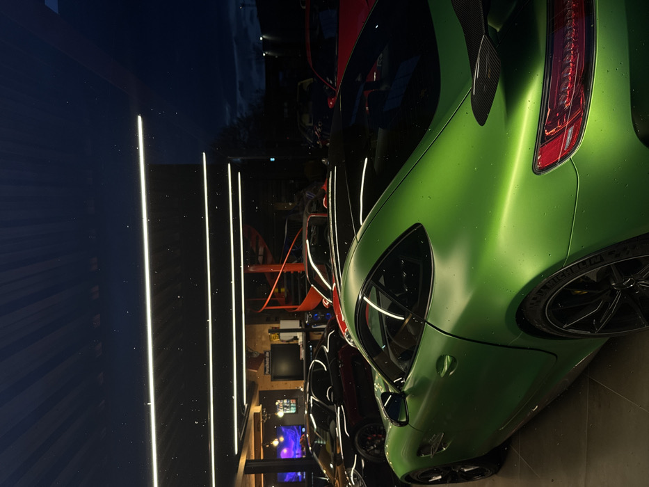 The image displays the interior of a car showroom or garage in Souffelweyersheim, France, during nighttime. In the foreground, a matte green Mercedes-AMG GT R sports car is prominently featured, positioned facing slightly left and away from the viewer. Its surface is speckled with small water droplets. The vehicle has black multi-spoke wheels and a carbon fiber rear spoiler.
Within the illuminated interior, behind the green car, multiple other high-performance sports cars, predominantly black, dark grey, and red, are parked. The ceiling is equipped with several bright, horizontal LED strip lights. To the right in the background, a distinctive red spiral staircase ascends. A large monitor displaying "Porsche.de" is visible on a wall. Beyond the glass facade of the building, the exterior scene is dark, characteristic of night, with faint distant lights and silhouetted foliage against a dark blue-black sky. No individuals are present in the visible area.