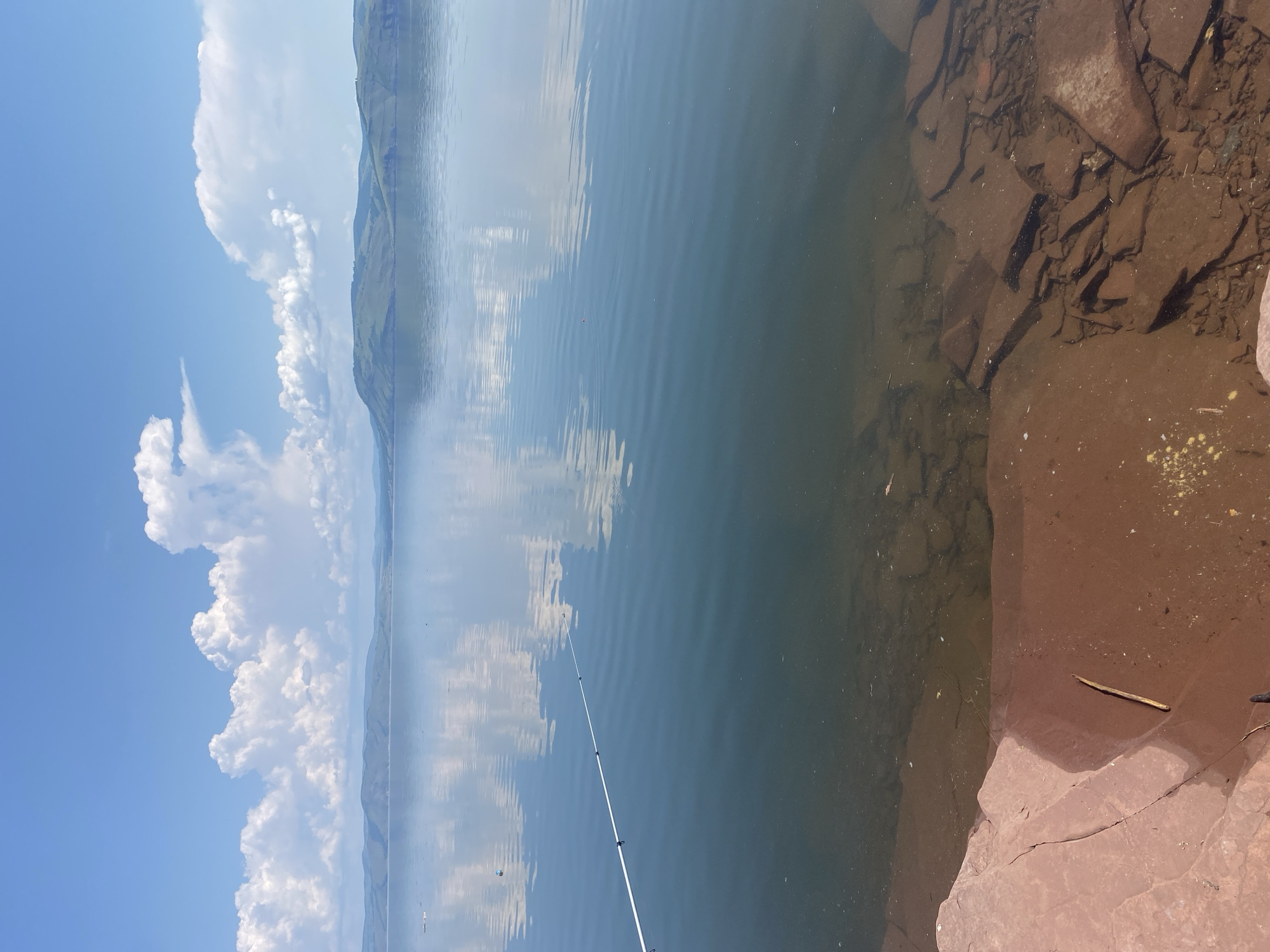 This image depicts a serene waterscape with a clear blue sky adorned with fluffy white clouds. The sky is reflected in the calm surface of a large body of water, creating a mirror-like effect. In the distance, rolling green hills form the horizon, their shapes softened by the atmospheric perspective. The foreground is comprised of rocky shorelines, with reddish-brown stones and submerged rocks visible in the shallow, clear water. A fishing rod extends into the water from the left side of the frame, suggesting a peaceful activity in progress. The scene evokes a sense of tranquility and natural beauty.
