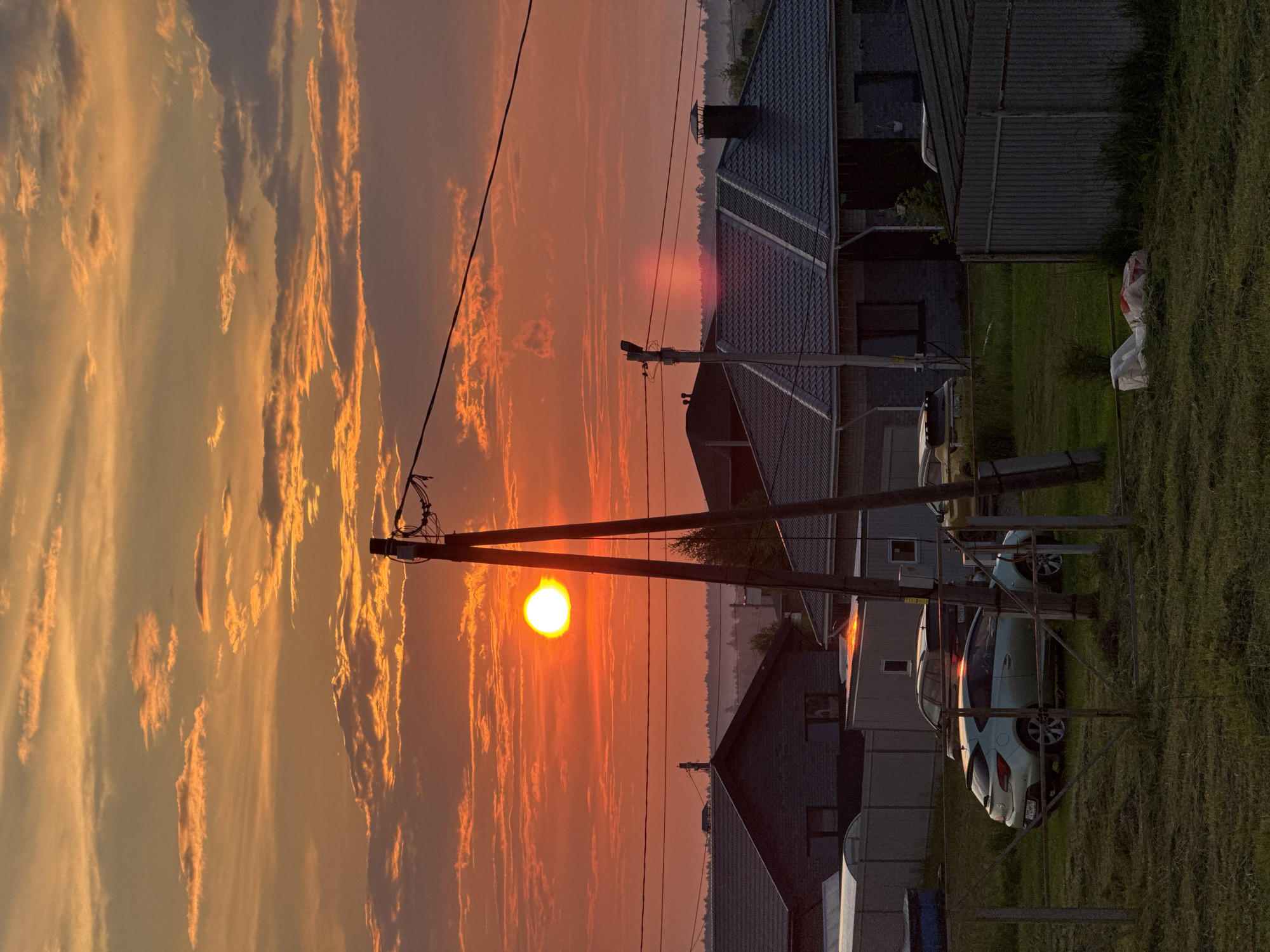 A low-angle, vertical-format, full-shot photograph depicts a sunset scene in the city of Izhevsk, Russia. The sky is filled with vibrant orange and yellow clouds, with a bright, full sun positioned slightly left of center. The sun is casting a bright orange glow across the sky. In the foreground, a tall, wooden utility pole with wires stretches from the bottom to the top of the frame, obscuring part of the sun and sky.
Below the sky, a row of houses with dark, tiled roofs is visible. The houses are a mix of modern designs with some appearing to be under construction. Several cars are parked in front of the houses, including a light-colored hatchback and a dark SUV. A metal fence runs along the right side of the frame, separating the houses from a grassy area in the foreground. The grass is dry and appears to have been recently cut. A pile of white bags and red material lies on the ground in the lower right quadrant of the image. The overall impression is of a rural or suburban setting at dusk, with a dramatic sunset providing a colorful backdrop.