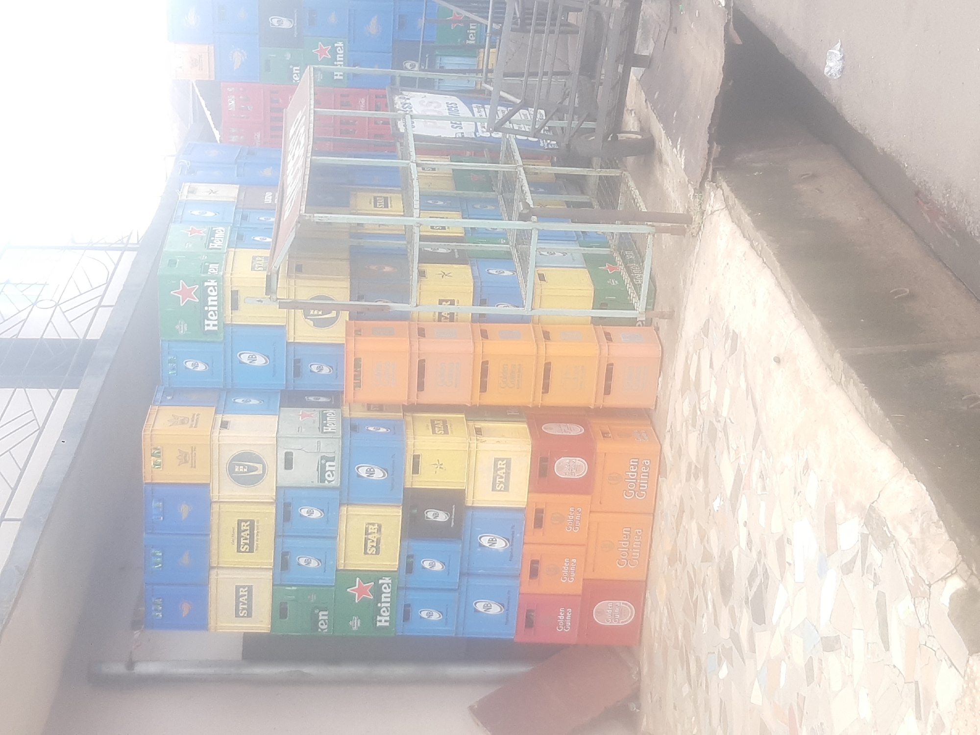 The image depicts a large stack of beverage crates, primarily beer crates, outside a building in Enugu, Nigeria. The crates are made of plastic and are in various colors including blue, yellow, green, and red. Many of the crates are labeled with brand names such as "STAR," "Heineken," and "Golden Guinea." Some of the crates appear to be empty, while others may contain bottles. The crates are stacked high, forming a wall-like structure. In the foreground, there is a mosaic tiled surface, possibly a sidewalk or patio, and a drainage ditch. Behind the crates, a metal shelving unit is visible, along with what appears to be a cart or trolley. The overall scene suggests a storage area or a point of sale for beverages. The lighting is bright, indicating it is daytime.