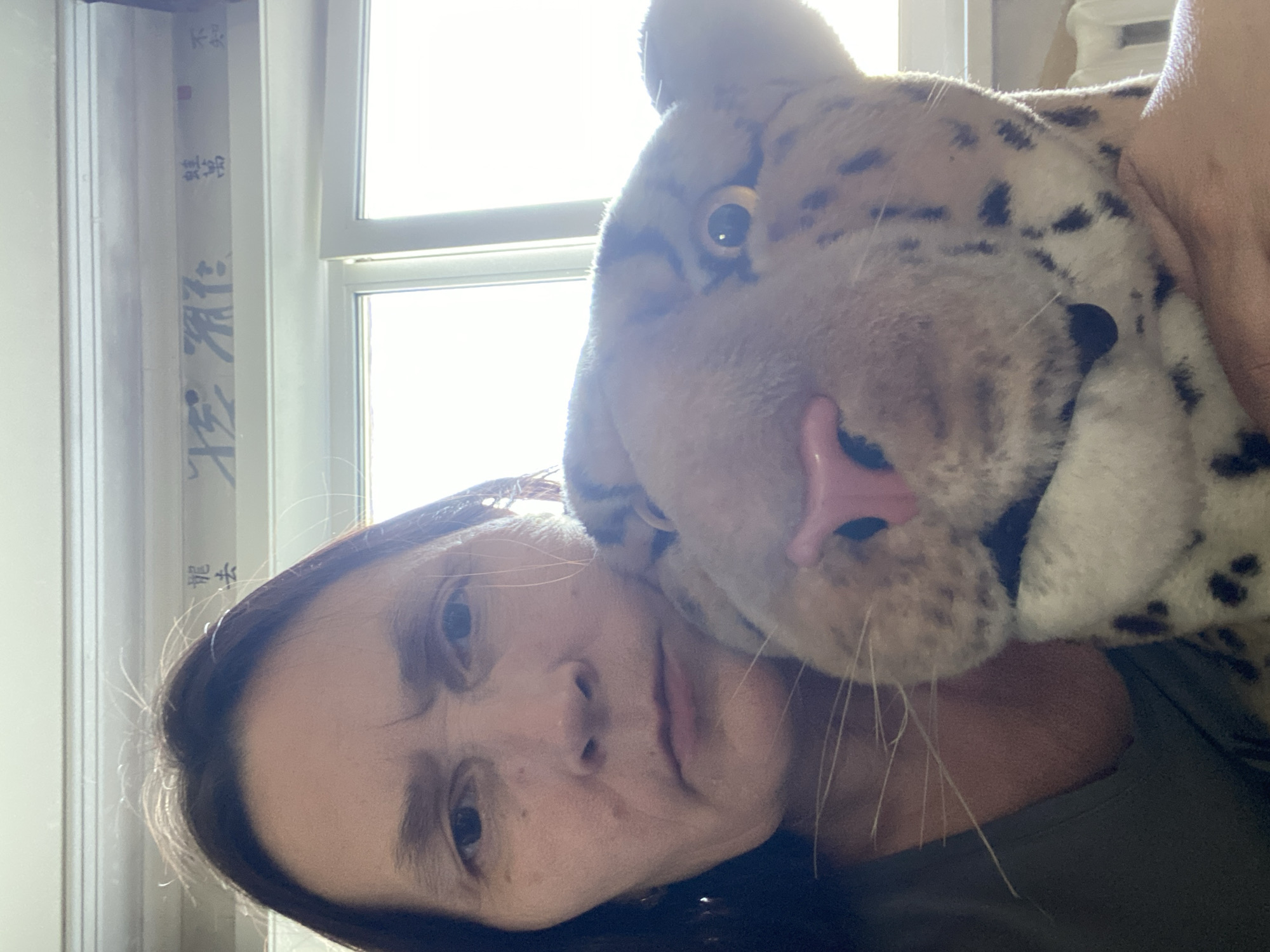 A close-up selfie shows a person holding a stuffed leopard. The person's face is partially visible, with brown hair and a neutral expression. The leopard's head is the dominant feature, with large eyes, a pink nose, and spotted fur. The background appears to be an indoor setting with a window and some text on a wall. There is no discernible information about the location in Novokuznetsk, Russia.