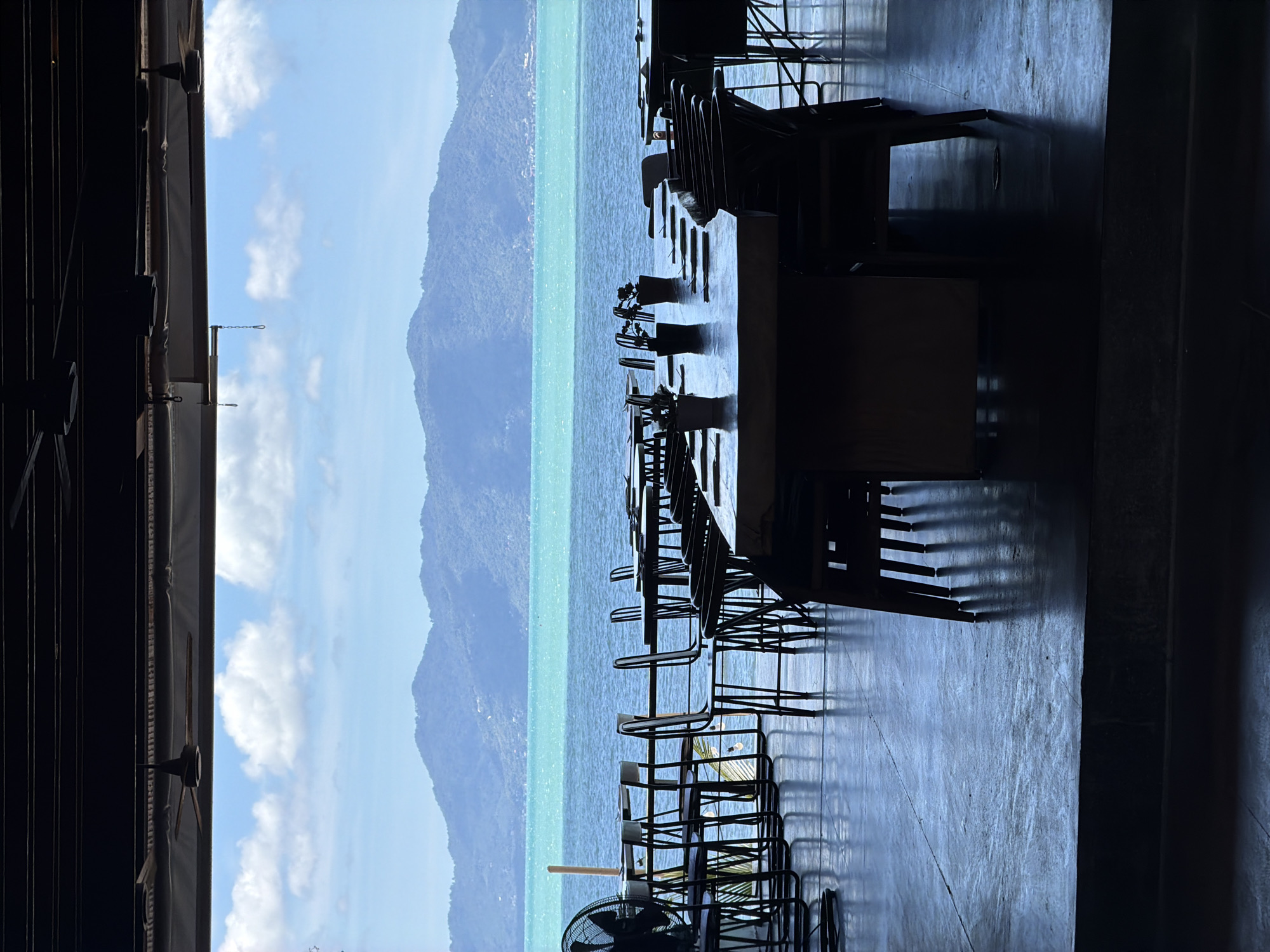 The image depicts a serene view of the ocean from a covered outdoor seating area. In the foreground, a long wooden table is set with placemats and small potted plants, surrounded by dark chairs. Further back, more tables and chairs are visible, some with chairs neatly tucked in. To the left, a large black fan stands near a row of tables. The scene opens up to a wide expanse of turquoise water, leading to a mountainous coastline under a bright, cloudy sky. The setting appears to be a restaurant or dining establishment overlooking the sea in Baan Bo Phut, Thailand. The lighting suggests it is daytime, with shadows cast on the floor from the furniture.