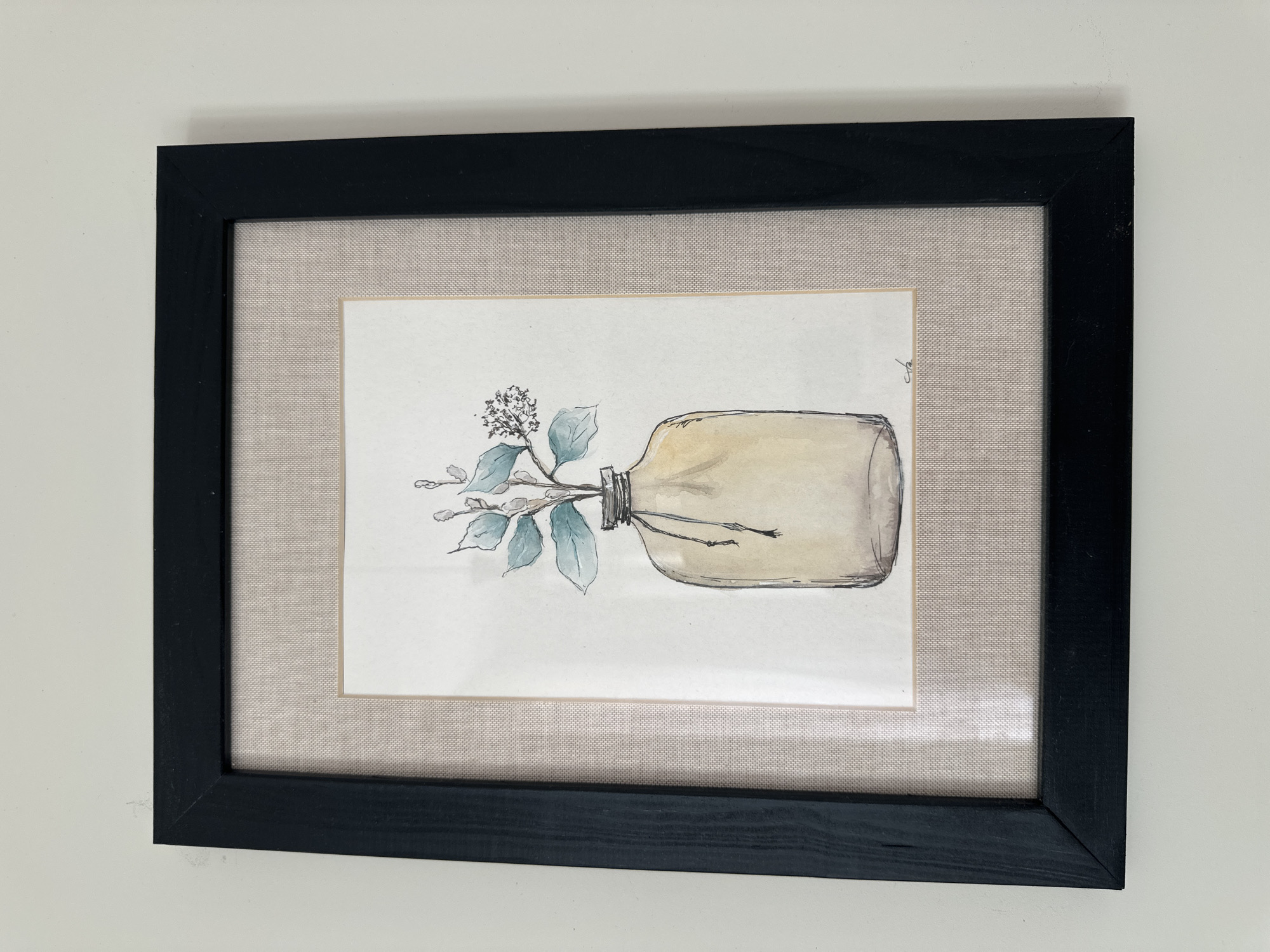 A framed watercolor painting of a glass bottle with a sprig of greenery is displayed on a white wall. The painting is matted with a beige fabric and framed in black wood. The bottle is depicted with a light brown hue and a string tied around its neck. The greenery features light blue leaves and small white flowers. The bottom right corner of the painting contains a small, illegible signature. The overall scene is minimalistic and appears to be an interior shot. The image was taken in Minsk, Belarus.