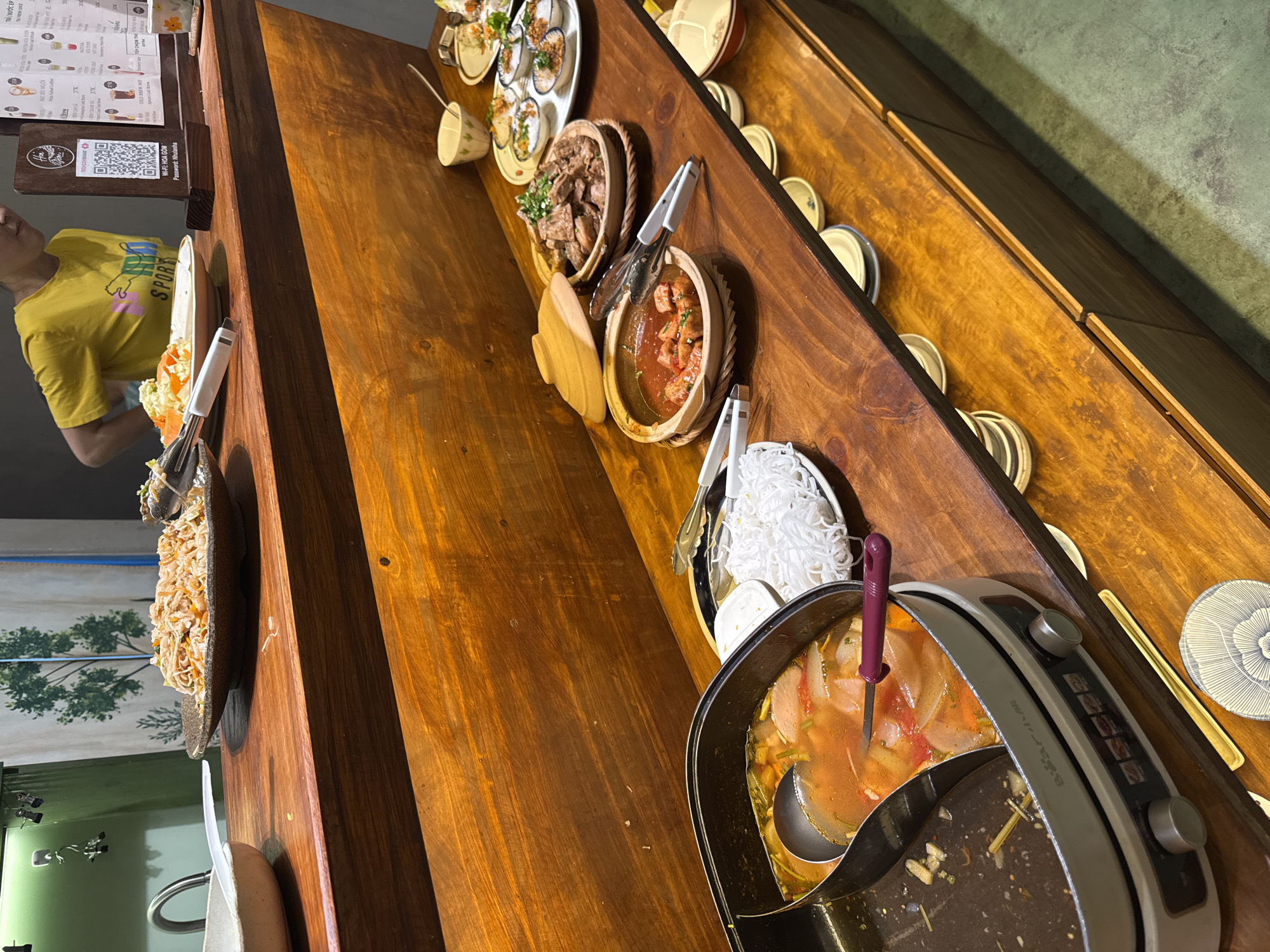 The image displays an indoor buffet or self-service food counter at a restaurant in Đà Nẵng, Vietnam, identified by a menu stand indicating "Wi-Fi: HOA GOM."
The scene is dominated by multi-tiered wooden counters. The upper counter, from left to right, features a white ceramic sink with a silver faucet, a white bowl with a plastic spoon, a large brown ceramic plate holding stir-fried noodles with serving tongs, and an adjacent plate of mixed vegetables with tongs. A person wearing a yellow t-shirt with "PUMA SPORTS" is partially visible behind this counter, facing away from the camera. A wooden menu stand, featuring Vietnamese text, drink prices, a QR code, and Wi-Fi credentials (HOA GOM, password: Nhulana), is positioned near the person.
The lower, main buffet counter, also wooden, presents multiple dishes. On the far left, an electric hot pot, branded "Bear," contains two compartments; one holds a reddish-orange broth with vegetables and food items, with a large metal ladle and a purple-handled utensil immersed. The second compartment is mostly empty with food residue. Adjacent to the hot pot is a white plate piled with thin white rice noodles (*bún*). Moving right, a ceramic bowl with a lid contains a reddish stew with meat or tofu chunks, equipped with serving tongs. Next to it, a wooden bowl holds a dark brown meat dish garnished with green herbs, also with serving tongs. Further right, a series of small white ceramic dishes, each containing a portion of food (possibly *bánh bèo*), are arranged on a larger platter. Various empty ceramic bowls and plates are distributed along the counter for patrons.
The background wall behind the upper counter is light green, featuring a white silhouette graphic of a tree. The floor is a textured, light green concrete surface.